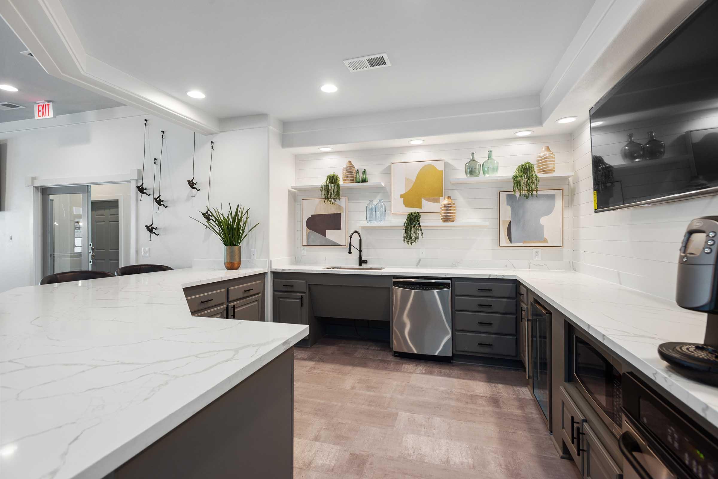 Modern kitchen featuring a large island with a white marble countertop, gray cabinets, and stainless steel appliances. Decorative shelves display colorful vases and artwork. Natural light floods the space, highlighting a clean, contemporary design with subtle greenery.