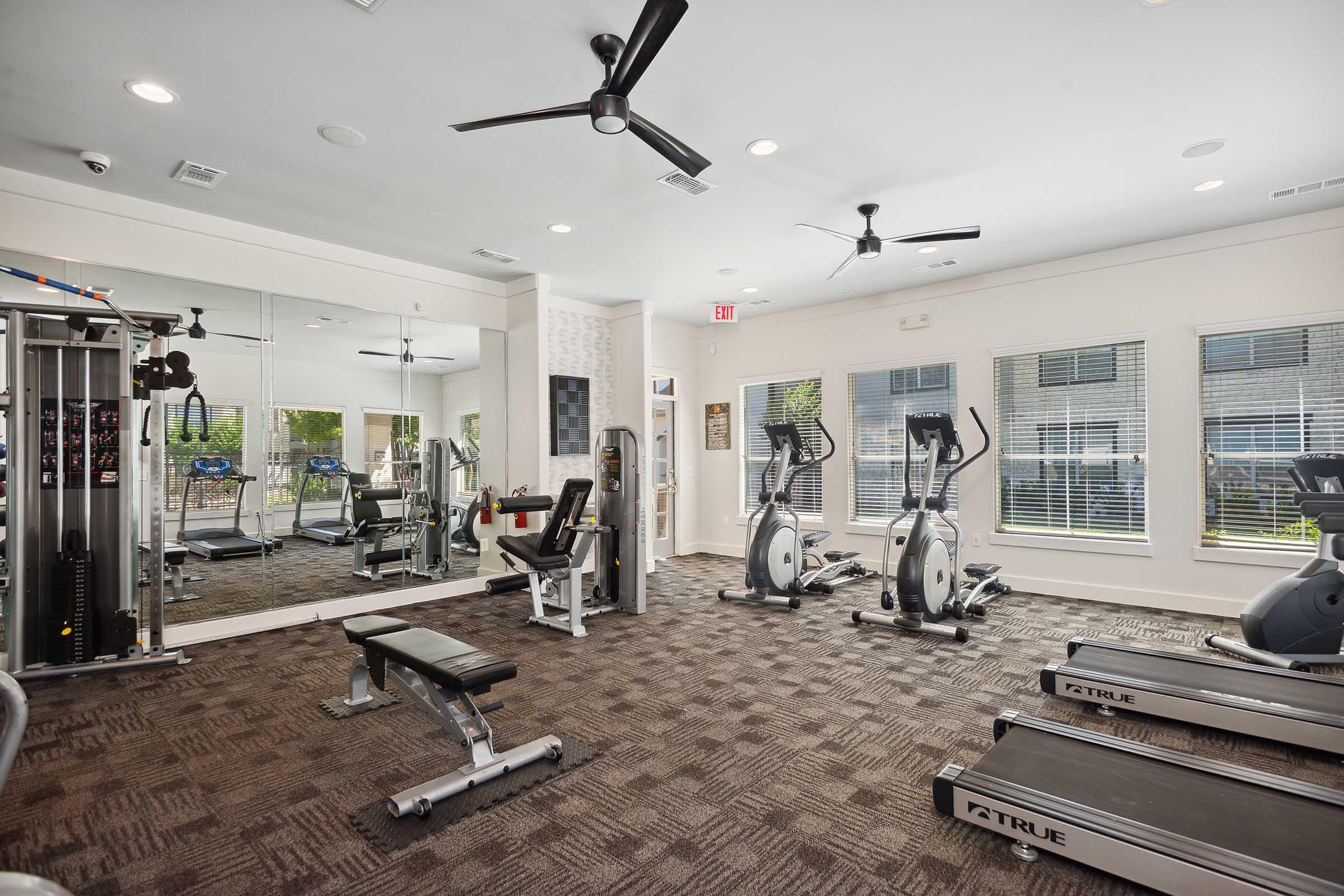 A modern gym area featuring exercise equipment including elliptical machines, a weight bench, and a multi-use cable machine. Large windows provide natural light, and there are ceiling fans for ventilation. A mirrored wall enhances the spacious feel of the room.