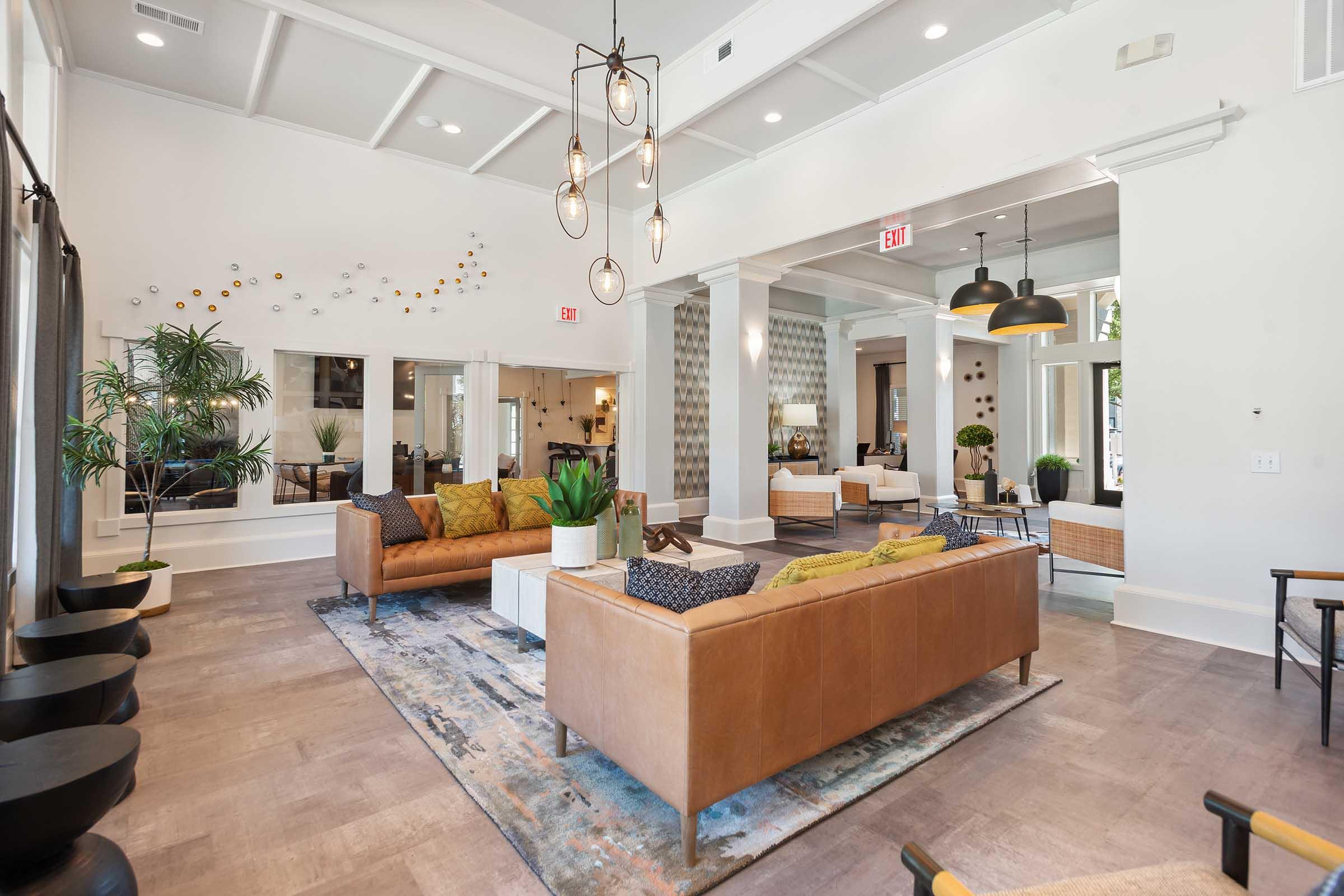 A modern, spacious lounge area featuring two stylish sofas, decorative plants, and contemporary light fixtures. The room has large windows, showcasing natural light, and stylish decor with a blend of earthy and vibrant colors. Seating arrangements provide a comfortable and inviting atmosphere.