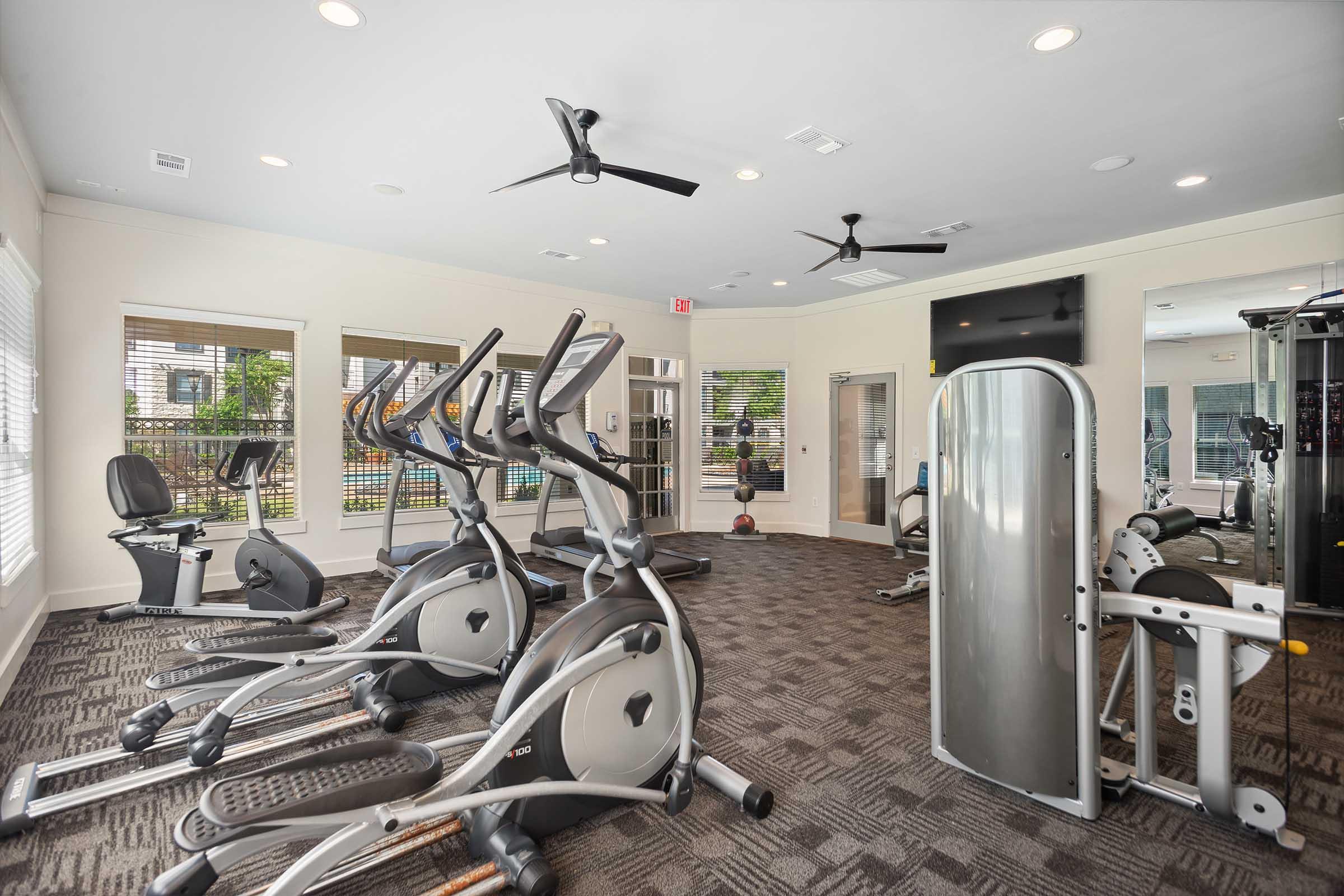 A modern gym interior featuring multiple elliptical machines, a stationary bike, and a multi-station strength training machine. The room is well-lit with ceiling fans and large windows, providing natural light and a view of the outdoors. The flooring is carpeted, creating a comfortable workout environment.