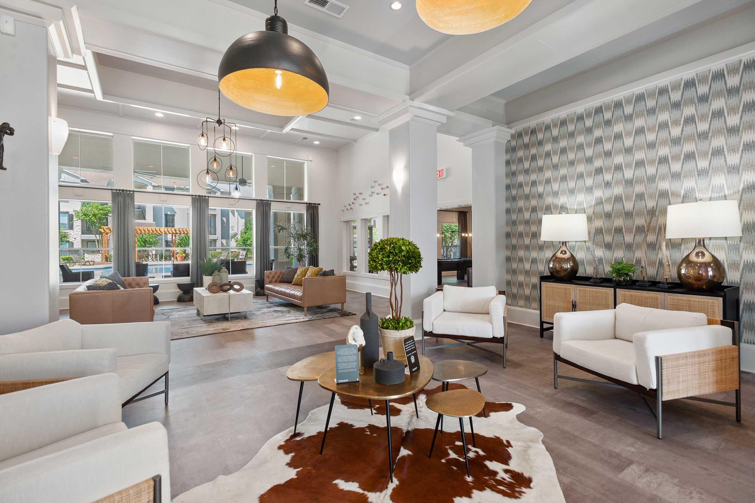 A modern and inviting lobby featuring stylish furniture, including white armchairs and a brown sofa. The space is well-lit with pendant lights, and has decorative elements like a cowhide rug, green plants, and a patterned wall. Large windows allow natural light, creating a warm atmosphere.
