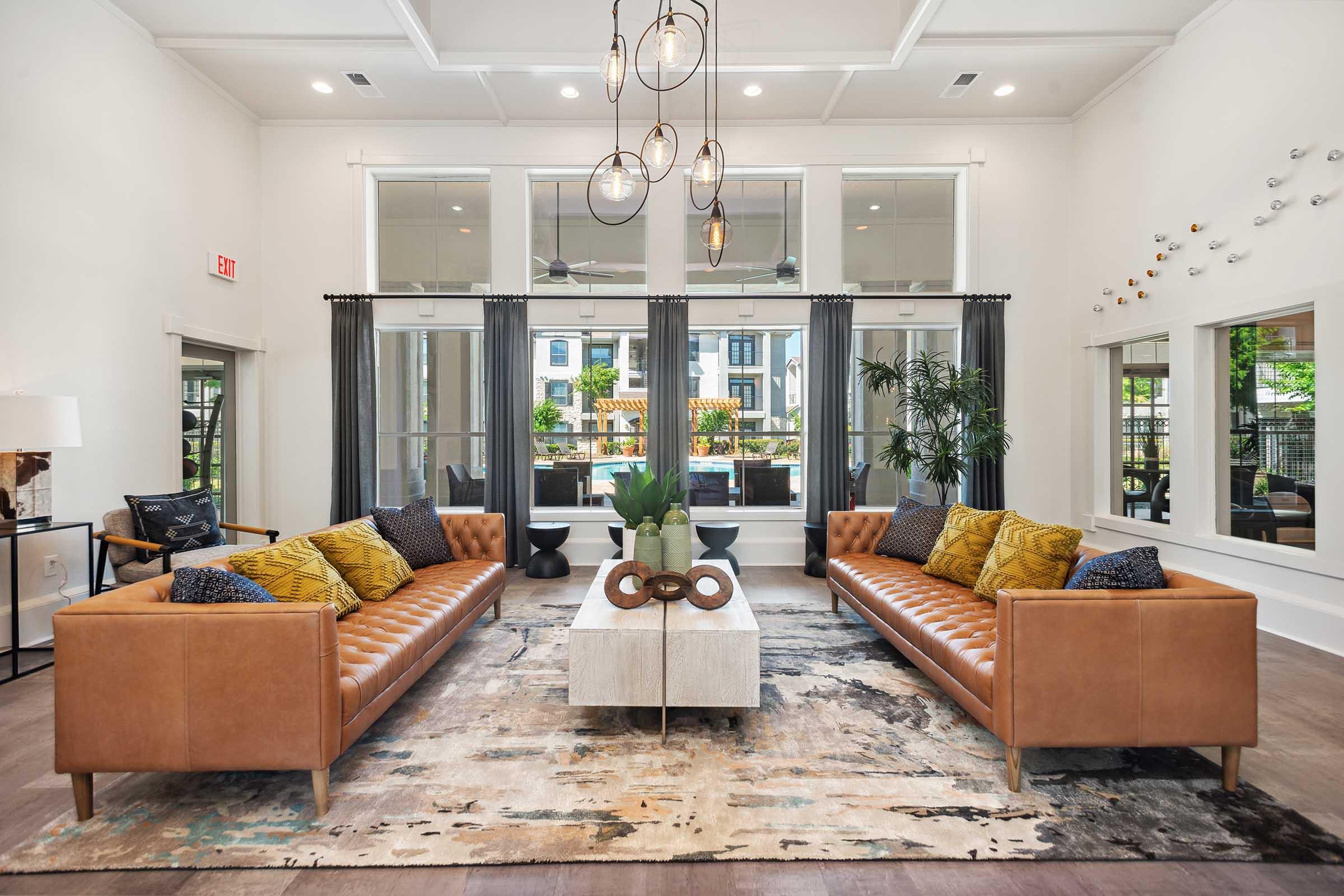 A spacious and modern living room featuring two brown leather sofas with yellow decorative pillows, a stylish coffee table, and large windows dressed with dark curtains. The decor includes plants and artistic wall features, creating a bright and inviting atmosphere.