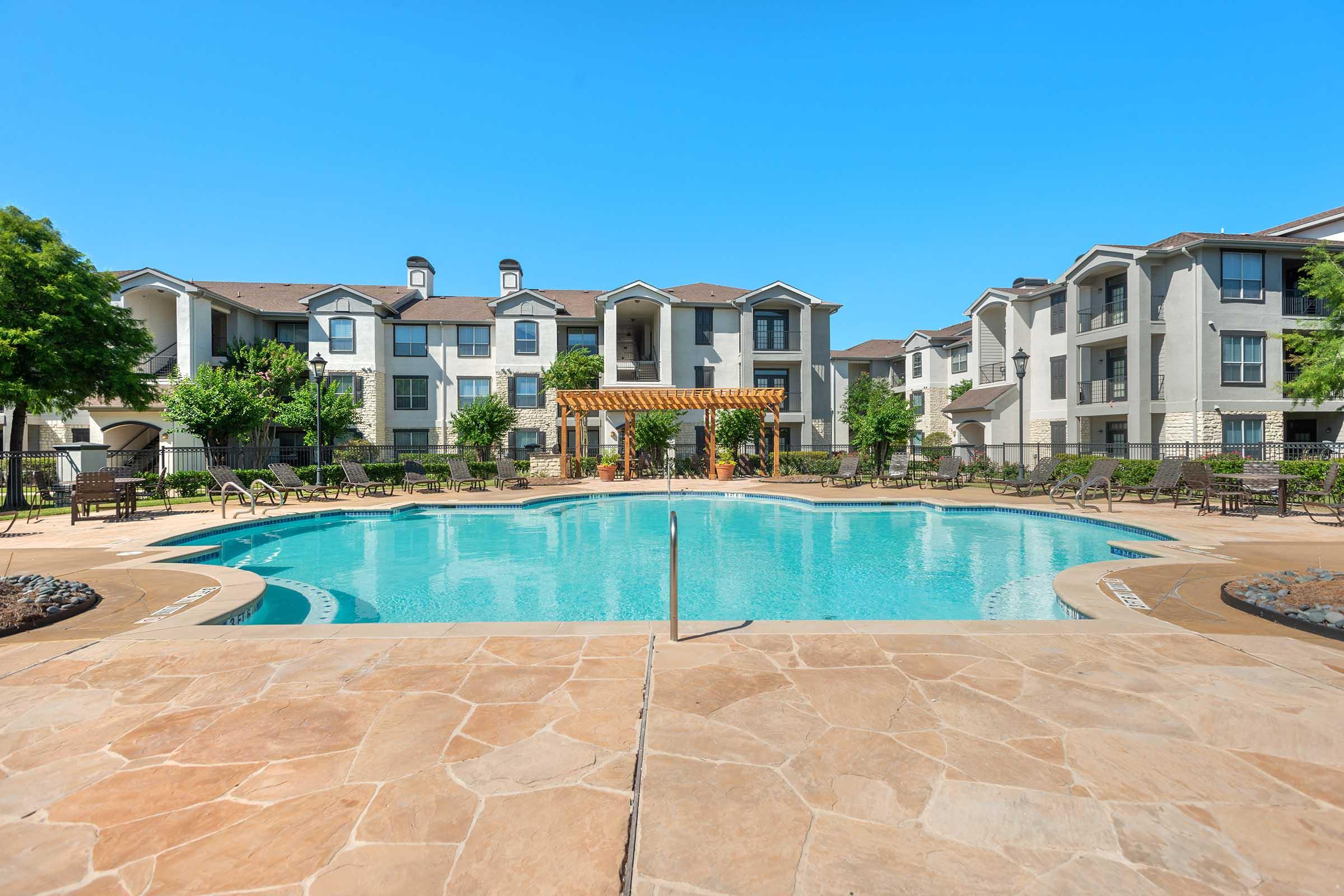 A clear blue swimming pool surrounded by landscaped areas and lounge chairs, with an apartment complex in the background featuring multiple stories and balconies. The sky is bright and sunny, creating a vibrant outdoor atmosphere.