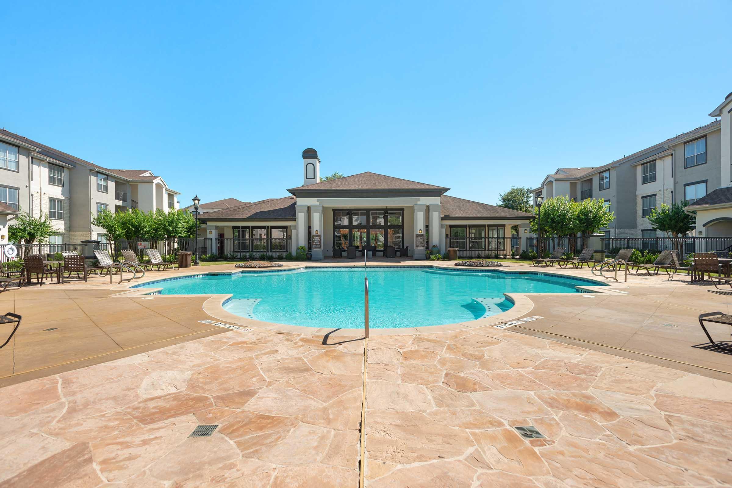 A clear blue swimming pool surrounded by a stone patio and lounge chairs. In the background, well-maintained apartment buildings and greenery are visible under a bright blue sky. The pool area features a central building with large windows.