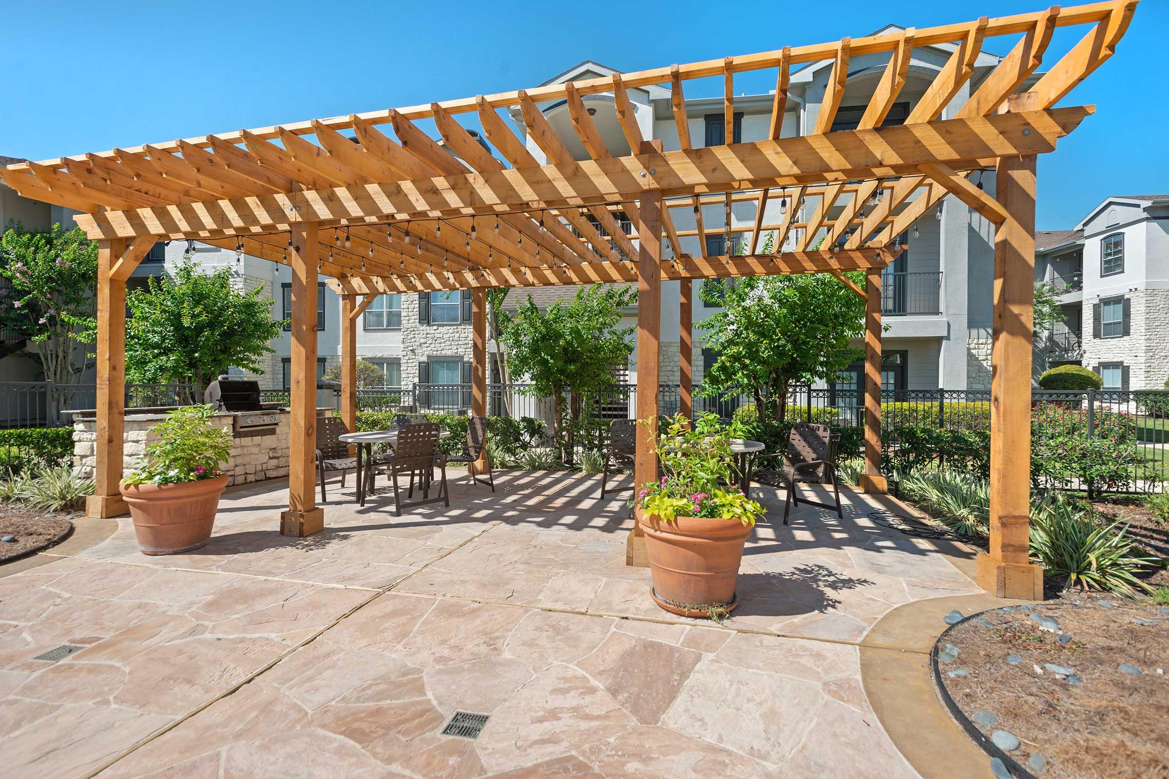 A wooden pergola with a slatted roof sits over a stone patio, surrounded by lush greenery and flowering plants in large pots. Outdoor furniture including a table and chairs is placed below the pergola, creating a serene space for relaxation. Nearby buildings are partially visible in the background under a clear blue sky.