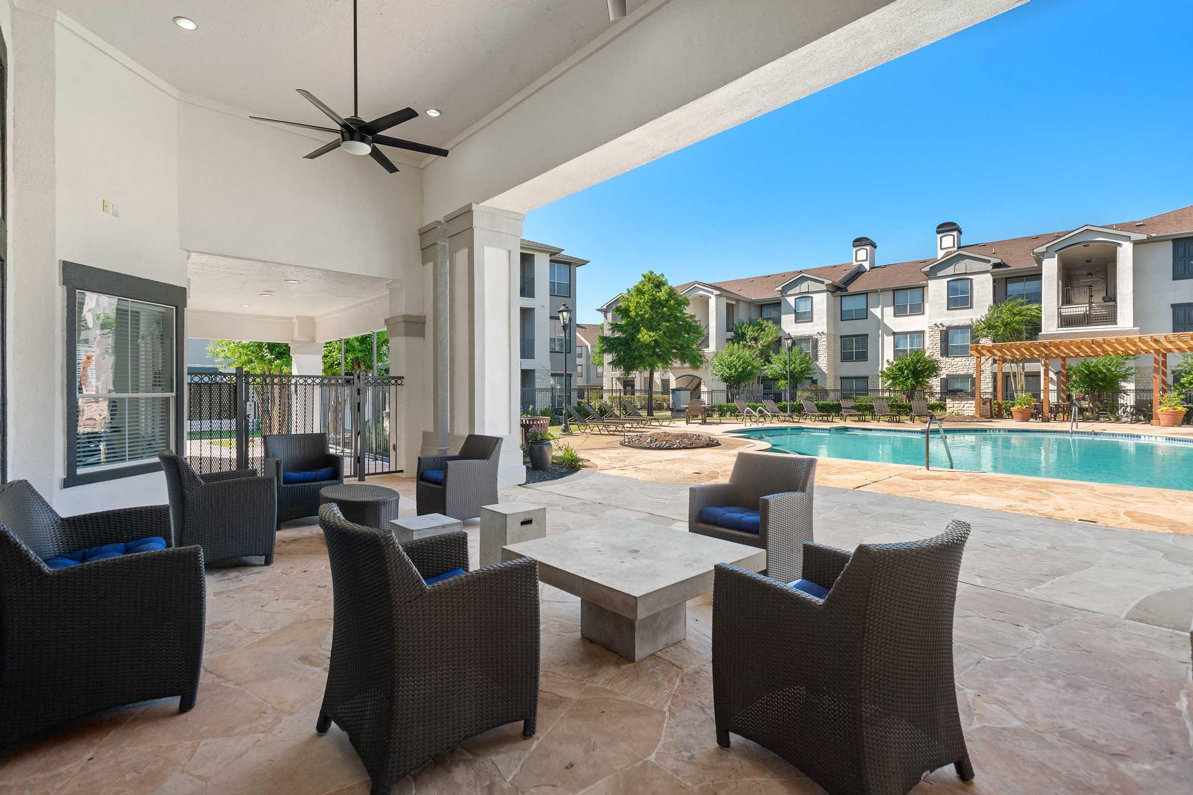 Outdoor seating area overlooking a swimming pool, with stylish wicker chairs and a modern table. Lush greenery and apartment buildings are visible in the background under a clear blue sky. The scene conveys a relaxing and inviting atmosphere.