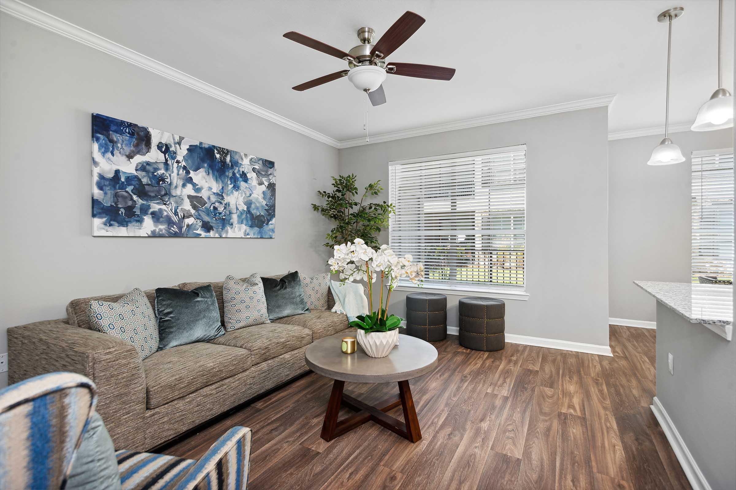 Living room featuring a cozy gray sofa adorned with decorative pillows, a round coffee table with a potted orchid centerpiece, and two modern stools. The walls are painted light gray, complemented by a blue abstract artwork above the sofa. Large windows provide natural light, enhancing the inviting atmosphere.