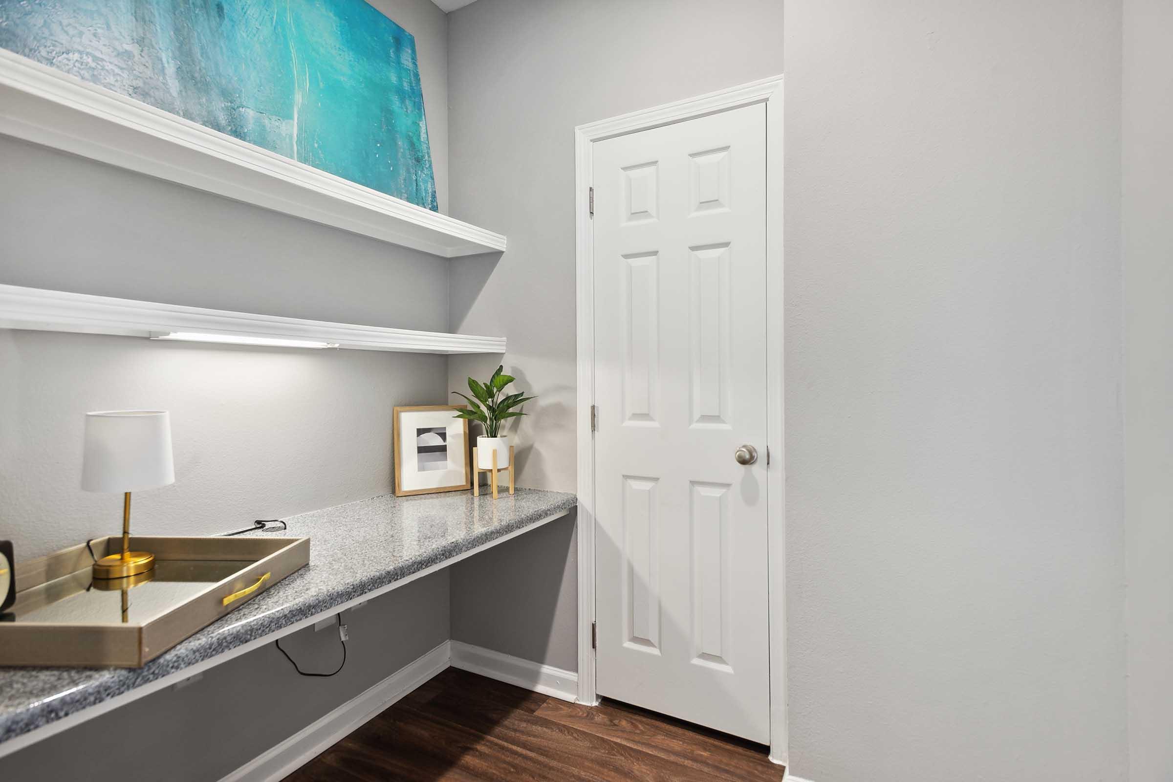 A modern, minimalistic workspace featuring a long countertop with a decorative tray, a small lamp, and a potted plant. Above the counter are two floating shelves that hold a framed picture and an abstract painting in blue tones. A white door is visible on the right, leading to another room.