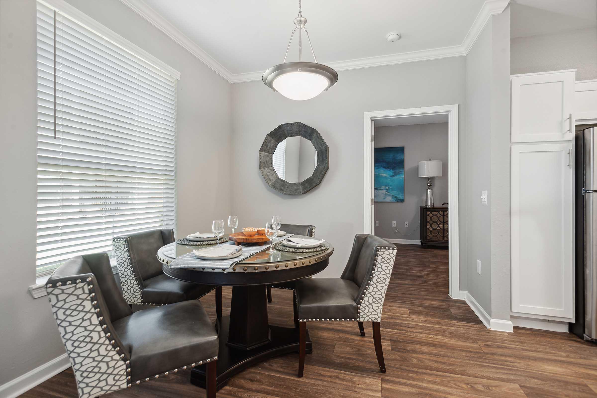 A modern dining area featuring a round table with four upholstered chairs. The table is set with plates and glassware. A decorative mirror is on the wall, and natural light streams in through a window with blinds. The floor is a warm wood tone, and a doorway leads into another room.