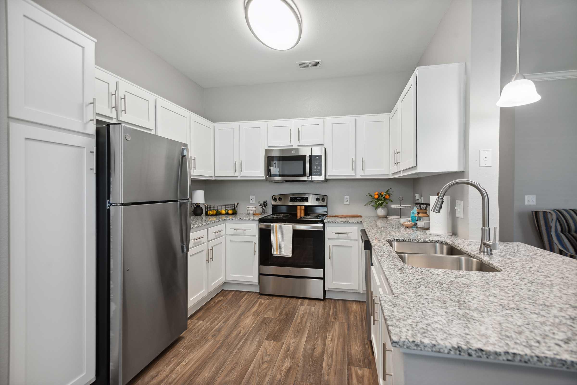 Modern kitchen with stainless steel appliances, including a refrigerator, microwave, and oven. White cabinets and a light granite countertop create a bright atmosphere. A sink is visible, and decorative items like flowers enhance the decor. Warm wood flooring complements the overall design.