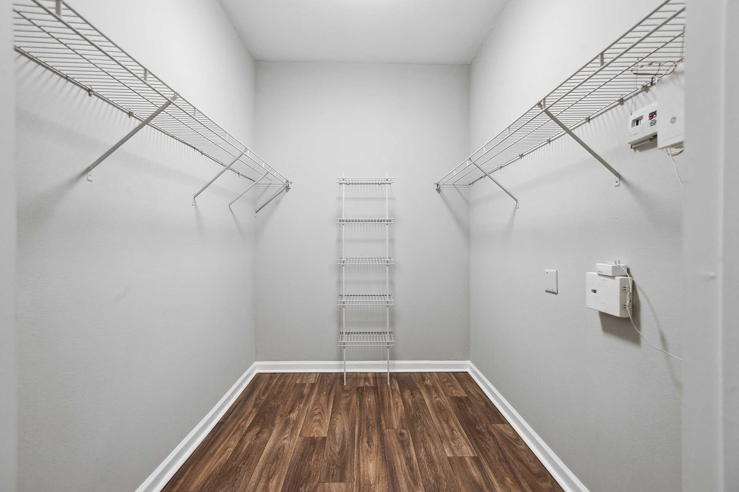 A minimalist closet featuring light gray walls, two wire shelves on either side, and a small wire shelf unit in the center. The floor is finished with laminate wood, giving it a warm appearance. There are no visible items stored in the closet, creating a spacious and empty feel.