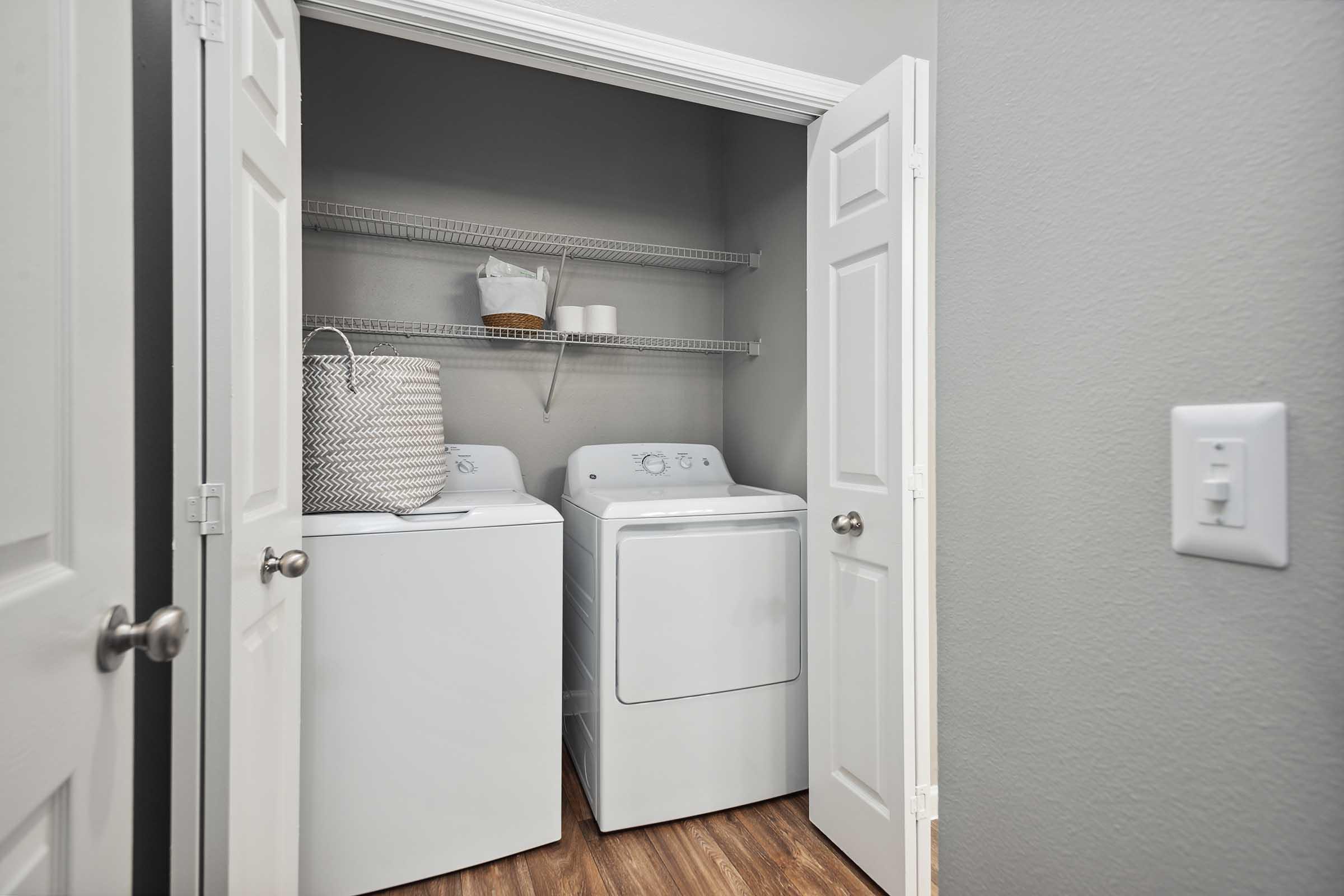 A laundry closet featuring a white washing machine and dryer side by side. There is a shelf above with a basket and some rolled towels. The closet has gray walls and white double doors, with wooden flooring visible. A light switch is on the right side of the image.