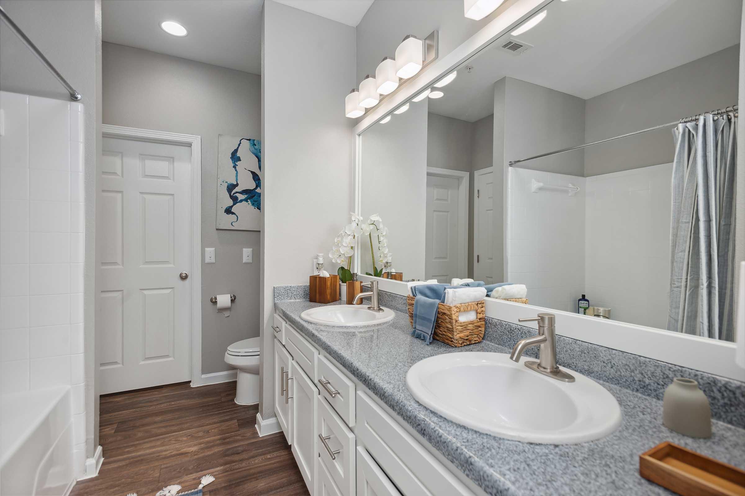 Modern bathroom with dual sinks, a large mirror, and stylish fixtures. The countertop is adorned with decorative items and towels. A shower area is visible, with a curtain partially drawn. The floor features wood-like tiles, and there’s a door leading to another room. Overall, the space is bright and contemporary.