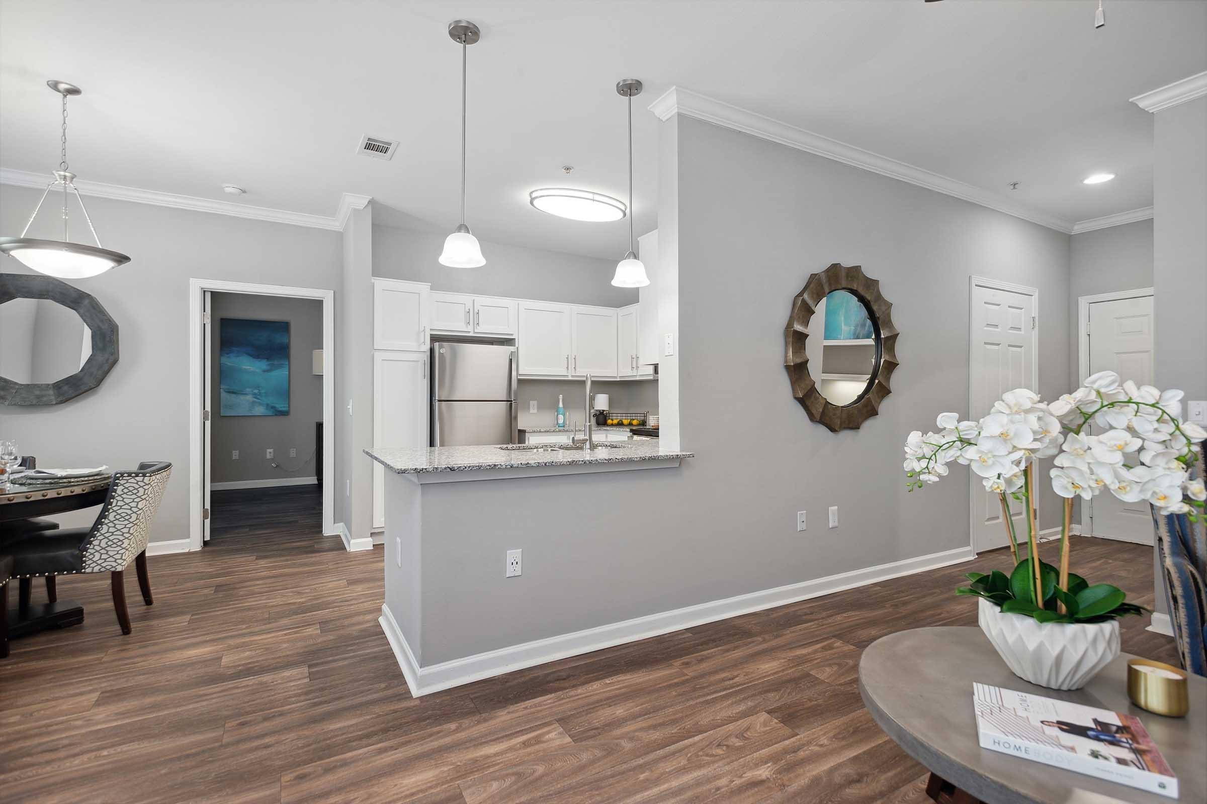 A modern, open-concept living space featuring a kitchen with stainless steel appliances and a granite countertop. There's a dining area with a round table set for four, a decorative mirror on the wall, and a small table with a plant and magazines. The walls are painted in light gray, creating a bright and inviting atmosphere.