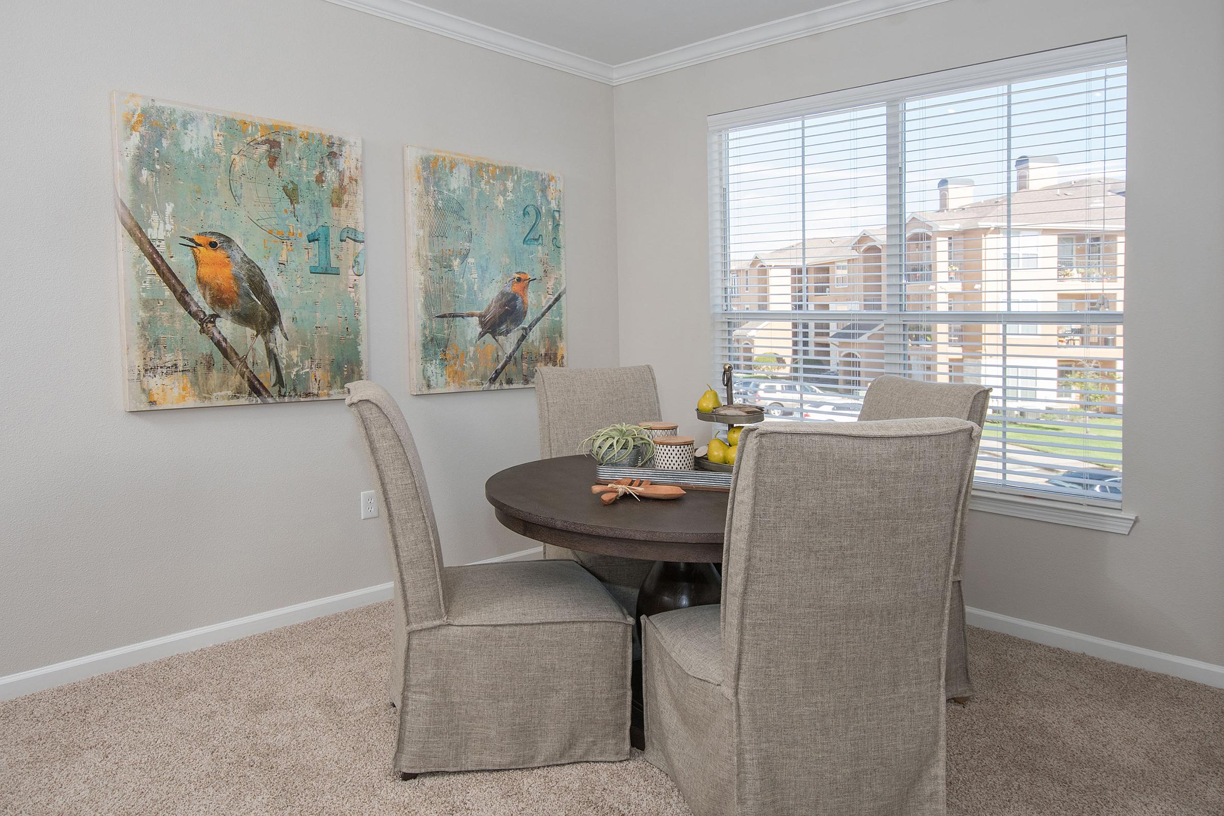 A cozy dining area featuring a round table surrounded by four upholstered chairs. Two framed bird-themed artworks hang on the wall, and a window with blinds lets in natural light. The room has a neutral color palette with carpet flooring and a view of an outdoor space.