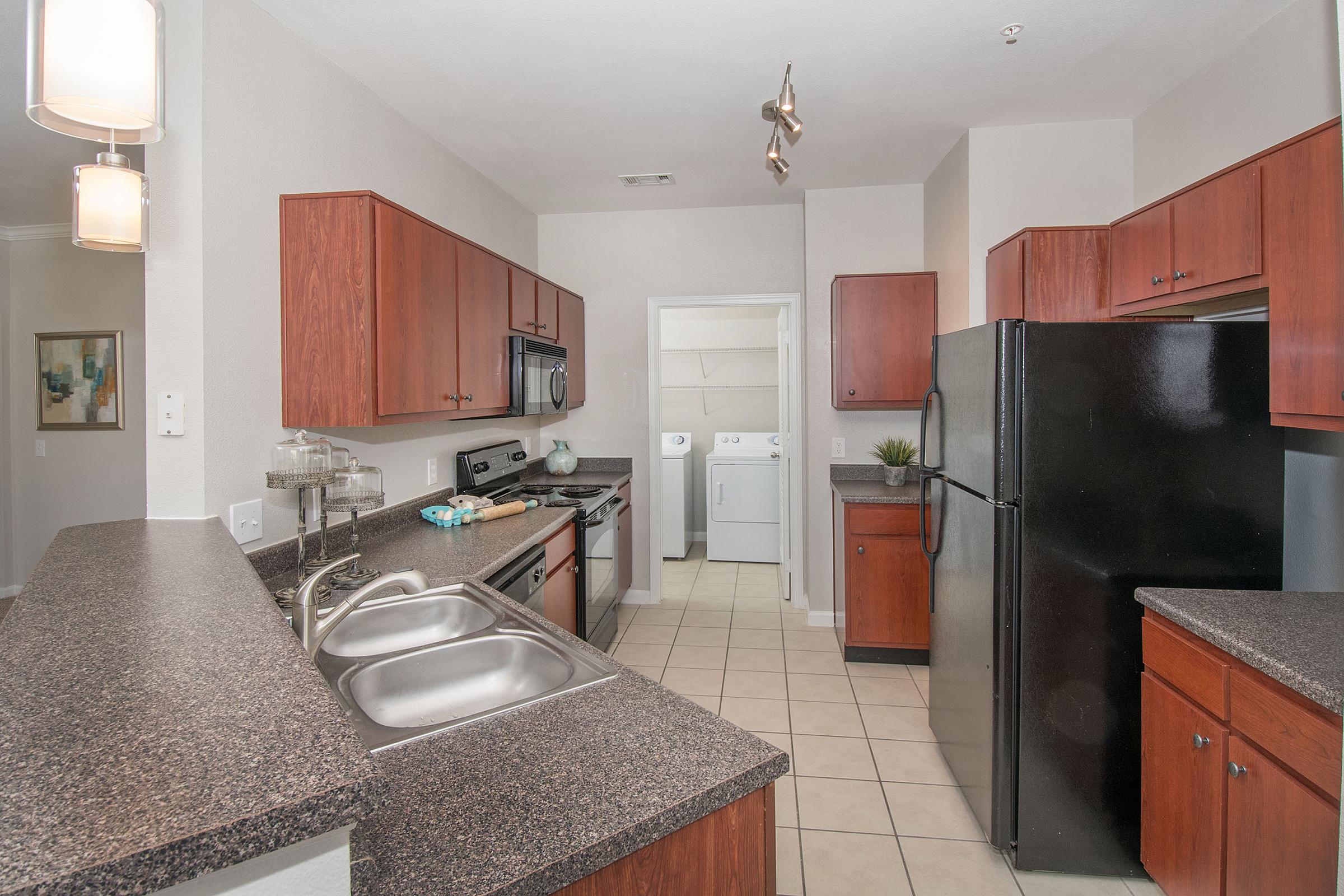 Modern kitchen featuring dark wood cabinets, stainless steel appliances, and granite countertops. A double sink is positioned under a window, and an island with bar seating is present. The adjoining area shows a laundry space accessible through a doorway. Neutral-colored walls and tile flooring complete the design.
