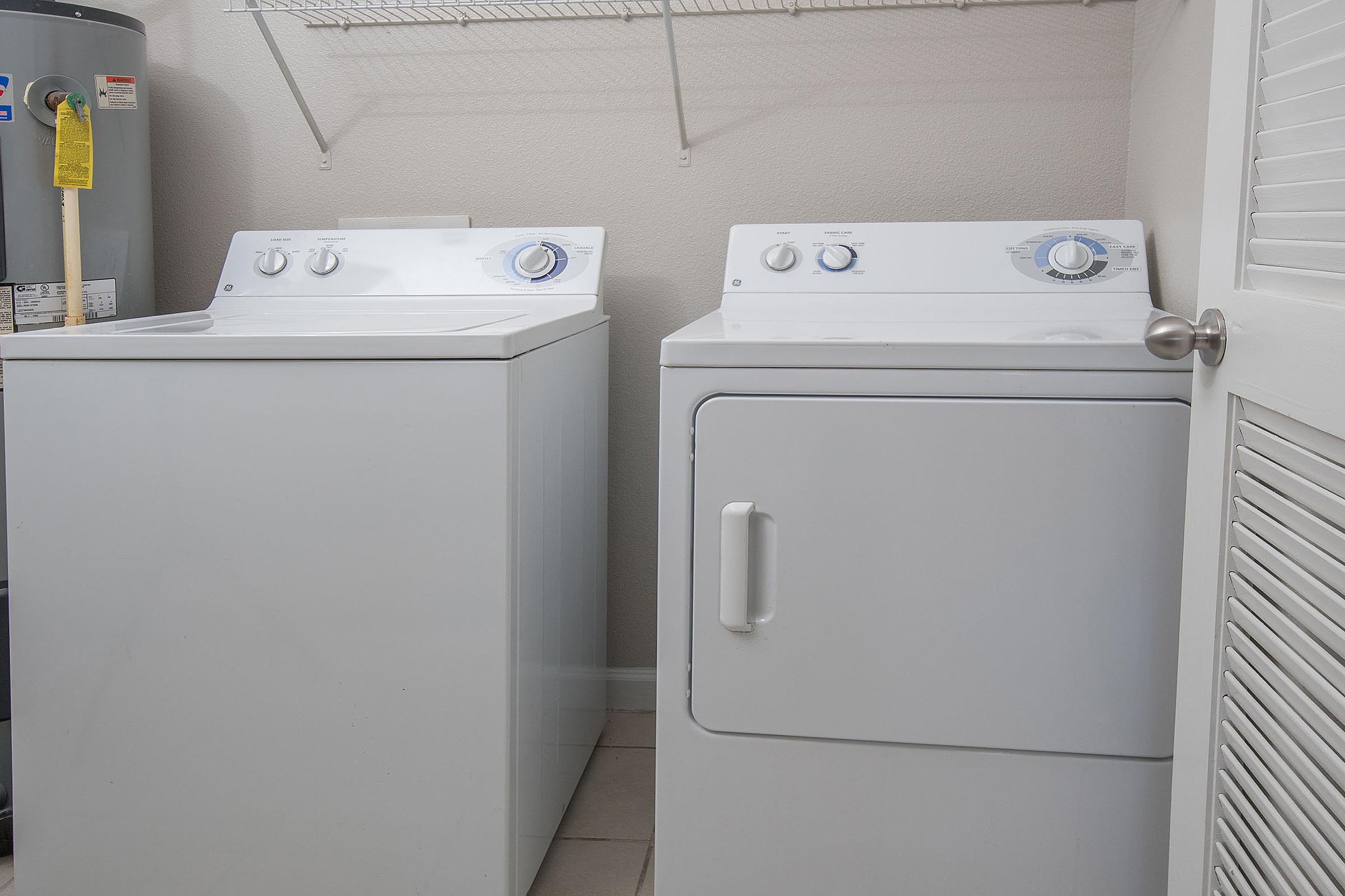 Two white appliances in a laundry room: a top-loading washing machine on the left and a front-loading dryer on the right. Both have dials and buttons for settings. The wall is light-colored, and there's a storage shelf above. A water heater is visible in the background. The floor is tiled.