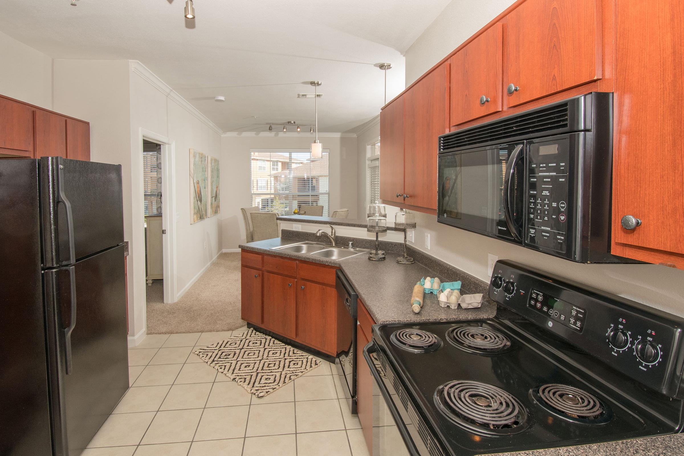 Modern kitchen featuring dark wood cabinets, a black refrigerator and stovetop, and a black microwave. The countertop is dark with a stylish rug on the tiled floor. Light floods the space from nearby windows, and a cozy living area is visible in the background.