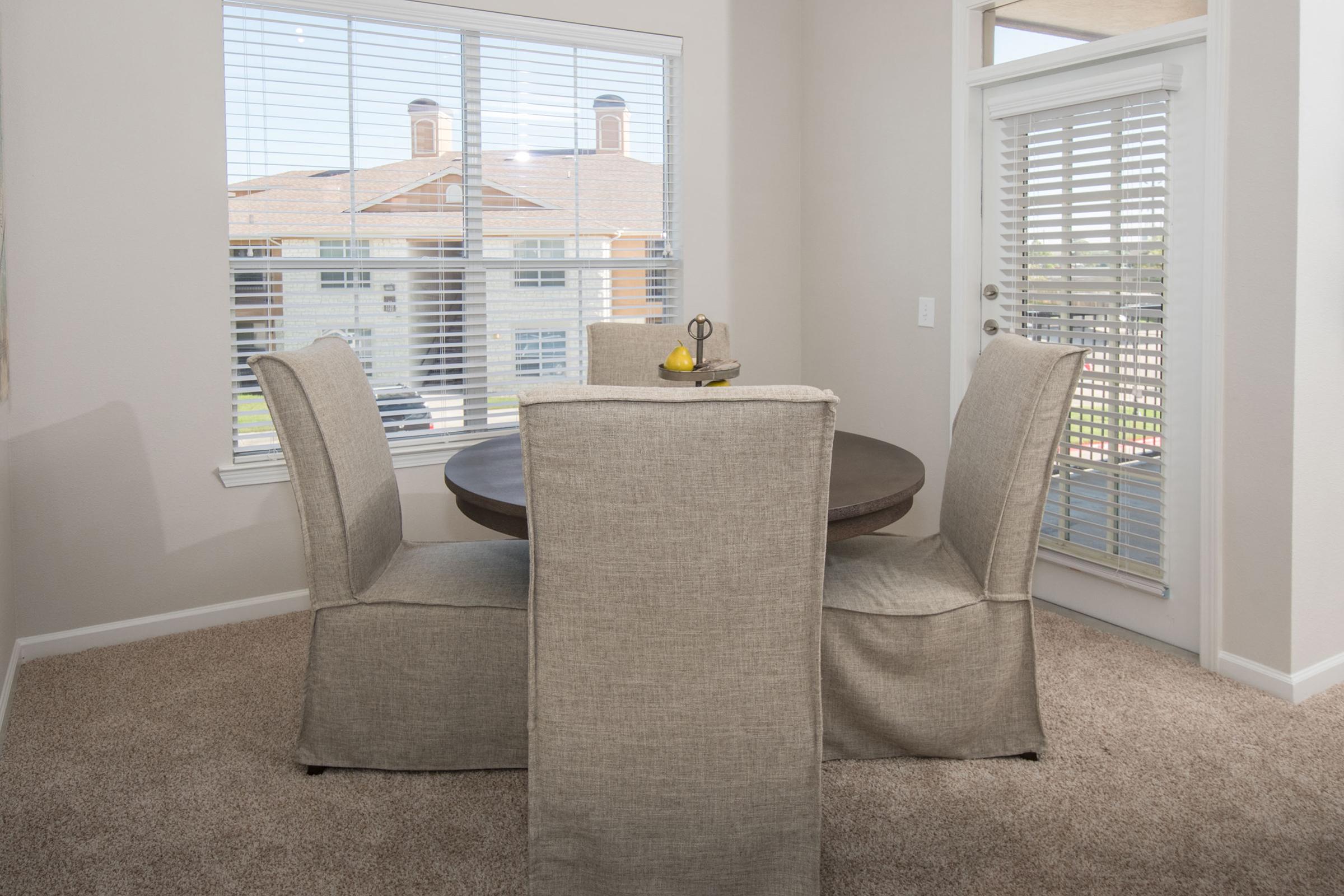 A cozy dining area featuring a round wooden table surrounded by four upholstered chairs. The room is well-lit by natural light streaming through large windows with white blinds, overlooking a neighborhood. A small decorative bowl sits on the table, adding a touch of elegance.