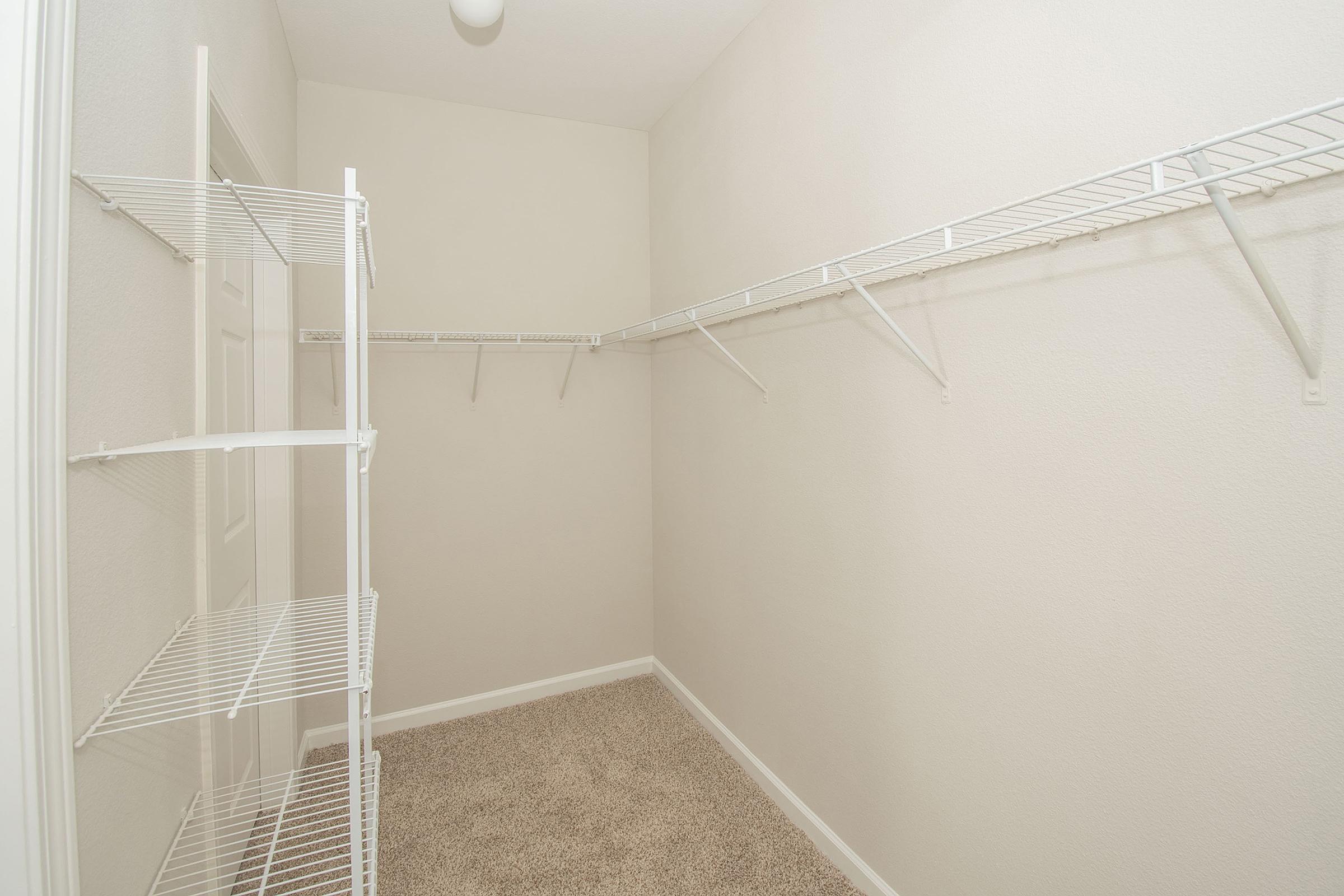 A spacious, empty closet featuring two wire shelving units on opposite walls. The closet has light-colored walls and a plush carpeted floor, with a ceiling light fixture providing illumination. The shelving offers storage options but currently has no items.