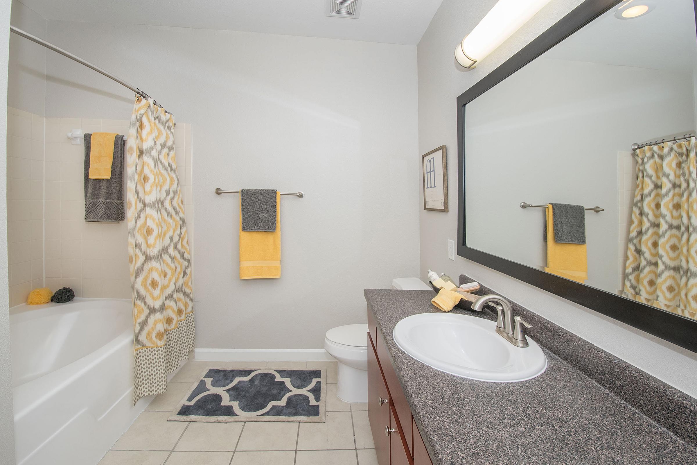 A modern bathroom featuring a bathtub with a shower curtain, sink with a dark countertop, and a large mirror. The décor includes gray, yellow, and white accents, with towels hanging on a towel rack and a rug on the floor. The walls are painted in a light color, enhancing the bright, clean atmosphere.