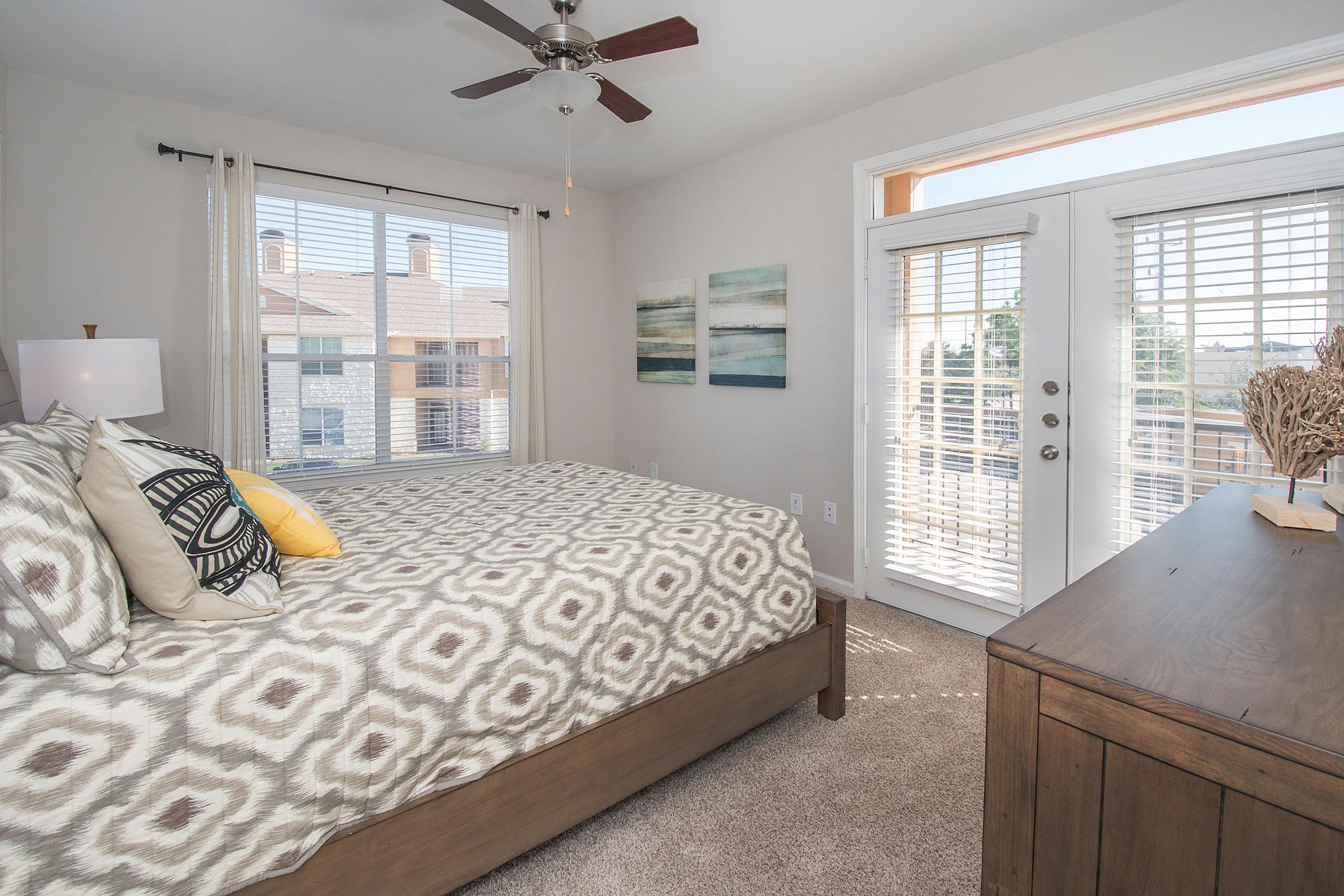 A cozy bedroom featuring a queen-sized bed with a patterned duvet and decorative pillows. The room has two large windows with blinds, a wooden dresser, and artwork on the wall, creating a light and inviting atmosphere. Access to a balcony is visible through glass doors.