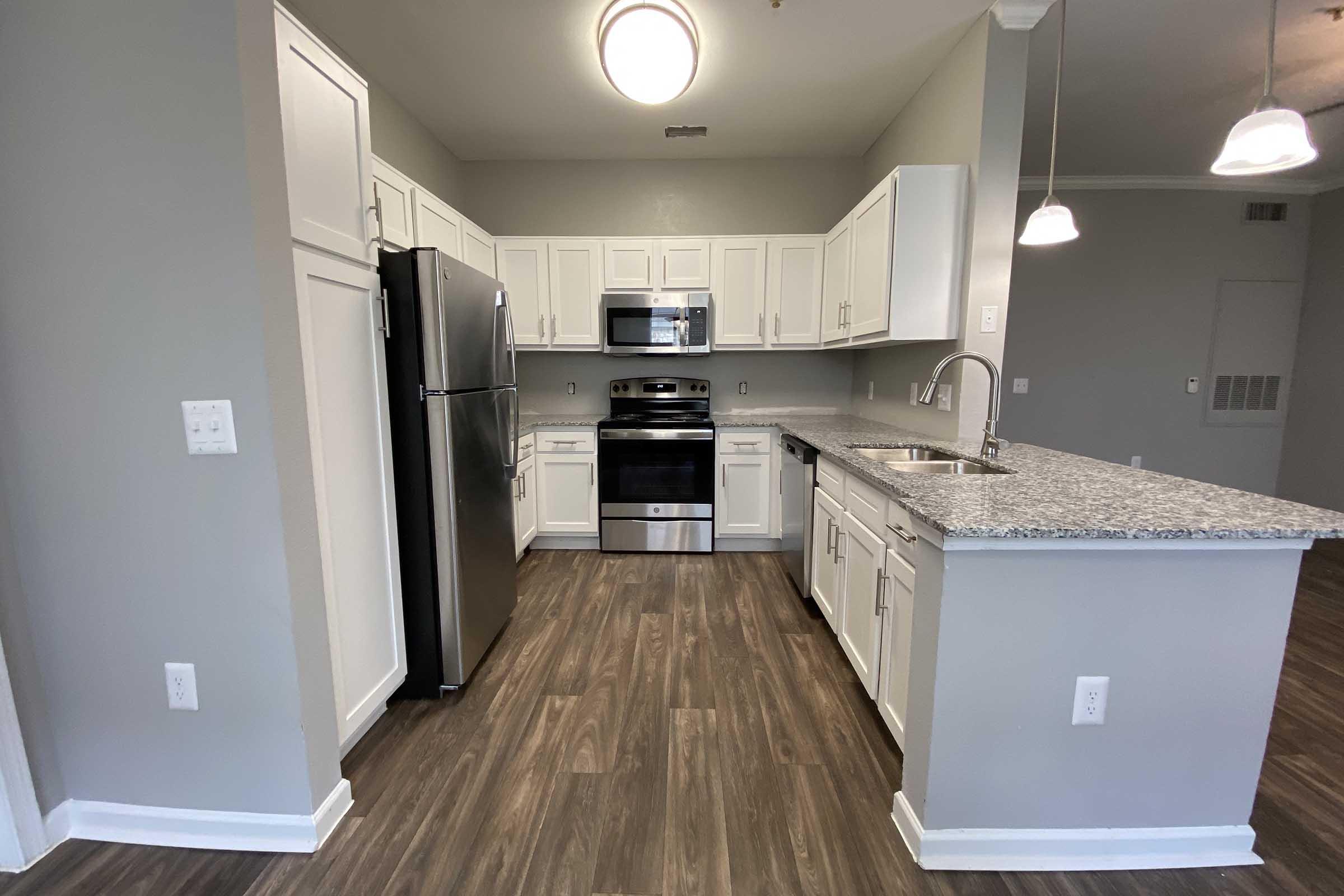 Modern kitchen featuring white cabinetry, stainless steel appliances, granite countertops, and a spacious layout. The flooring is dark wood, enhancing the contemporary look. Bright overhead lighting illuminates the area, creating a welcoming atmosphere.