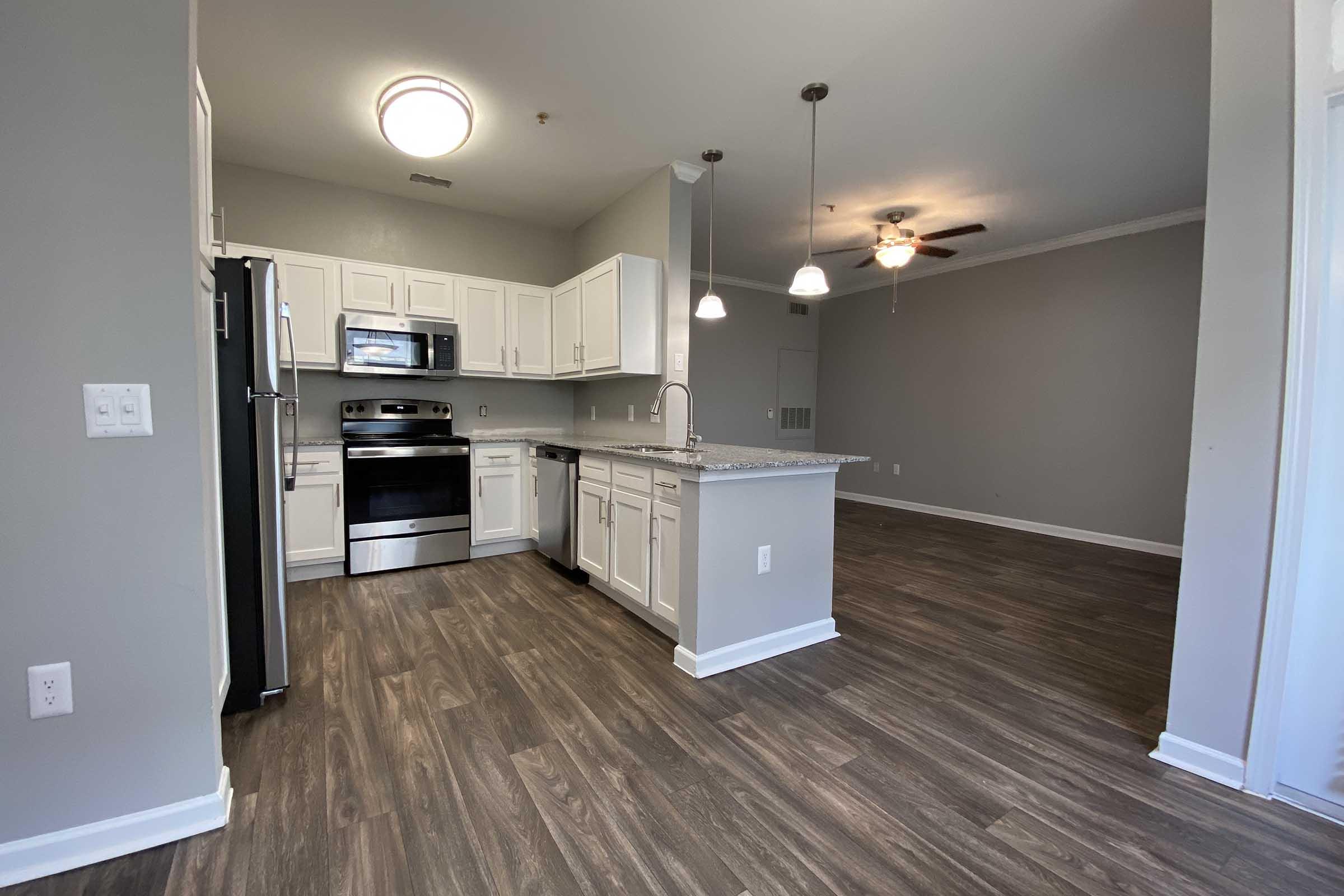 A modern kitchen featuring stainless steel appliances, white cabinets, and granite countertops. The open layout connects to a spacious living area with hardwood-style flooring and natural light from pendant and ceiling fixtures. The walls are painted in a neutral gray tone.