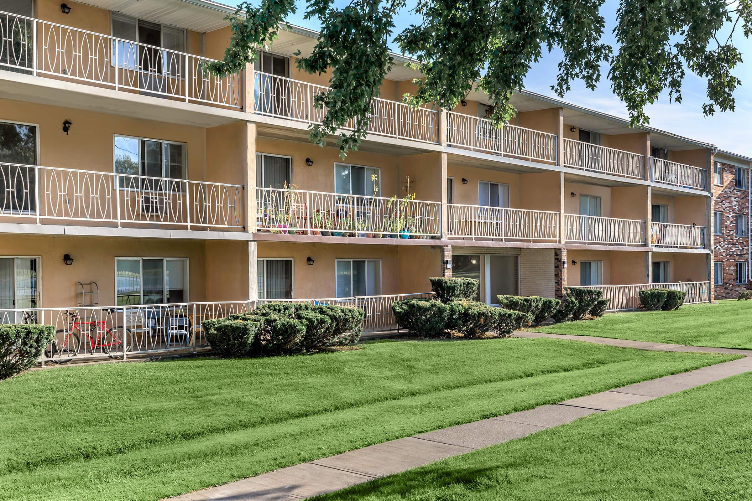 A view of a multi-story apartment building featuring beige exterior walls, white railings on the balconies, and large windows. The well-maintained green lawn in front has neatly trimmed bushes, and a paved walkway leads up to the building. The scene is bright and inviting, suggesting a peaceful residential environment.