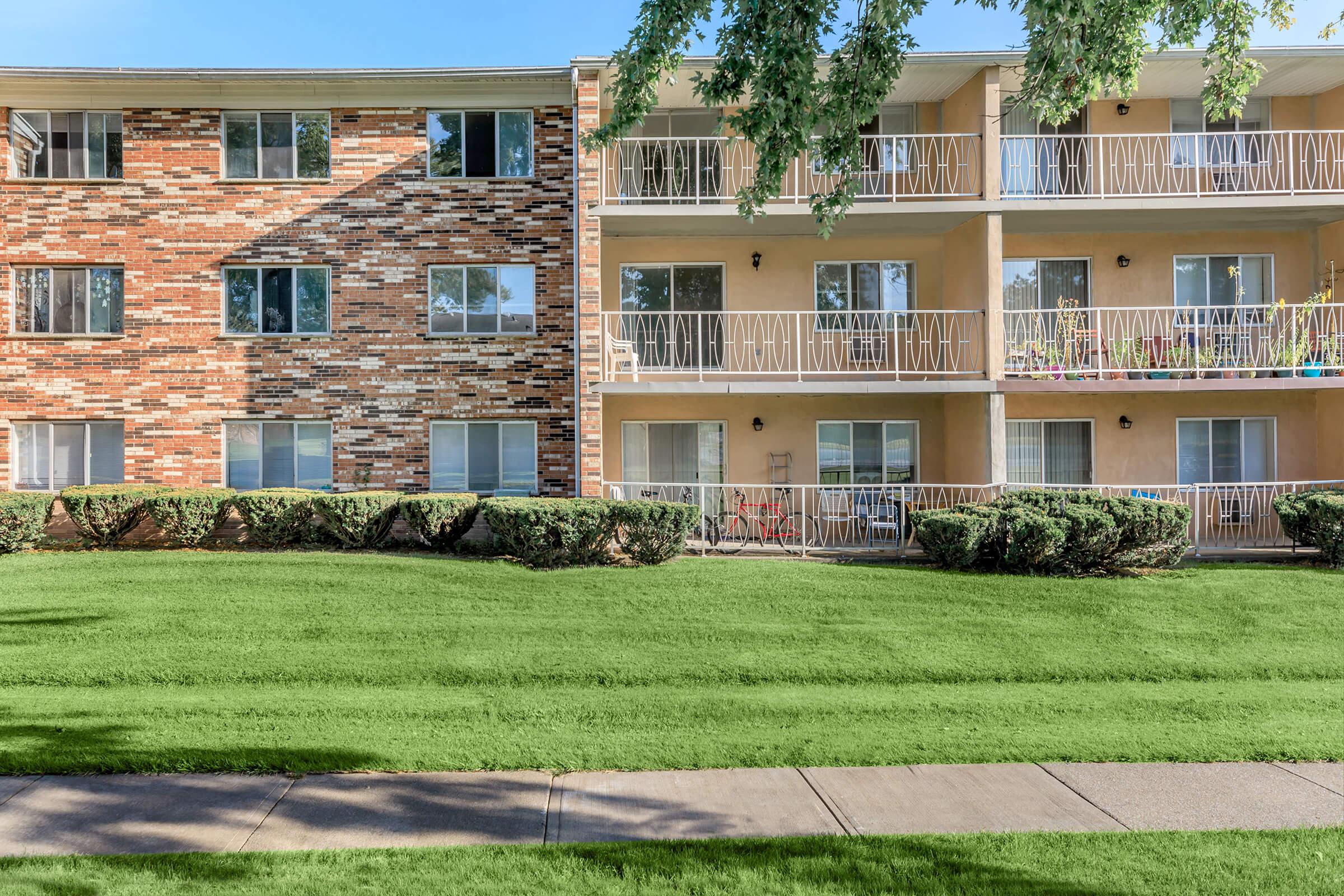 A view of two adjacent apartment buildings with a grassy area in front. One building features a brick facade with large windows, while the other has a lighter exterior and multiple balconies. The neatly maintained lawns and walkways enhance the residential setting.