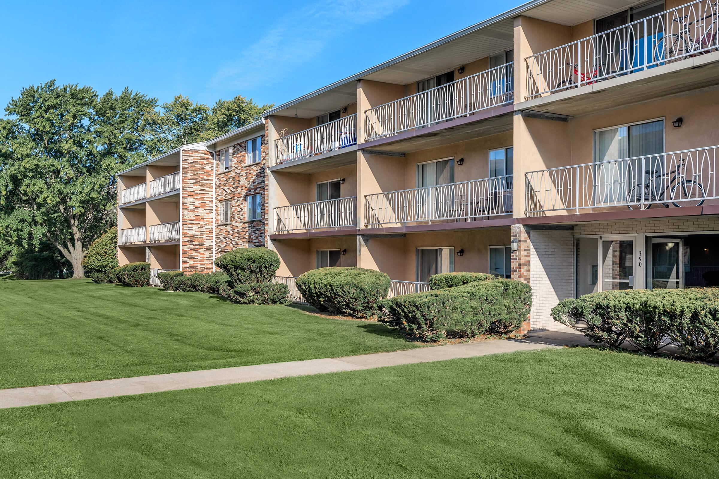 A three-story apartment building featuring balconies and a landscaped lawn with manicured shrubs. The building has a mix of brick and stucco exterior, with several windows visible. Clear blue skies and trees are visible in the background, creating a welcoming residential atmosphere.