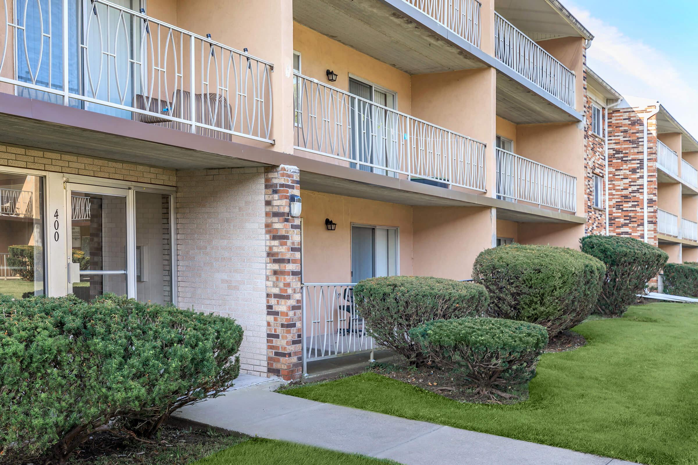 A view of a multi-story apartment building with balconies, featuring beige and brick exterior. The foreground includes neatly trimmed bushes and a sidewalk leading to the entrance, while the lawn is well-maintained, creating a tidy and inviting atmosphere.