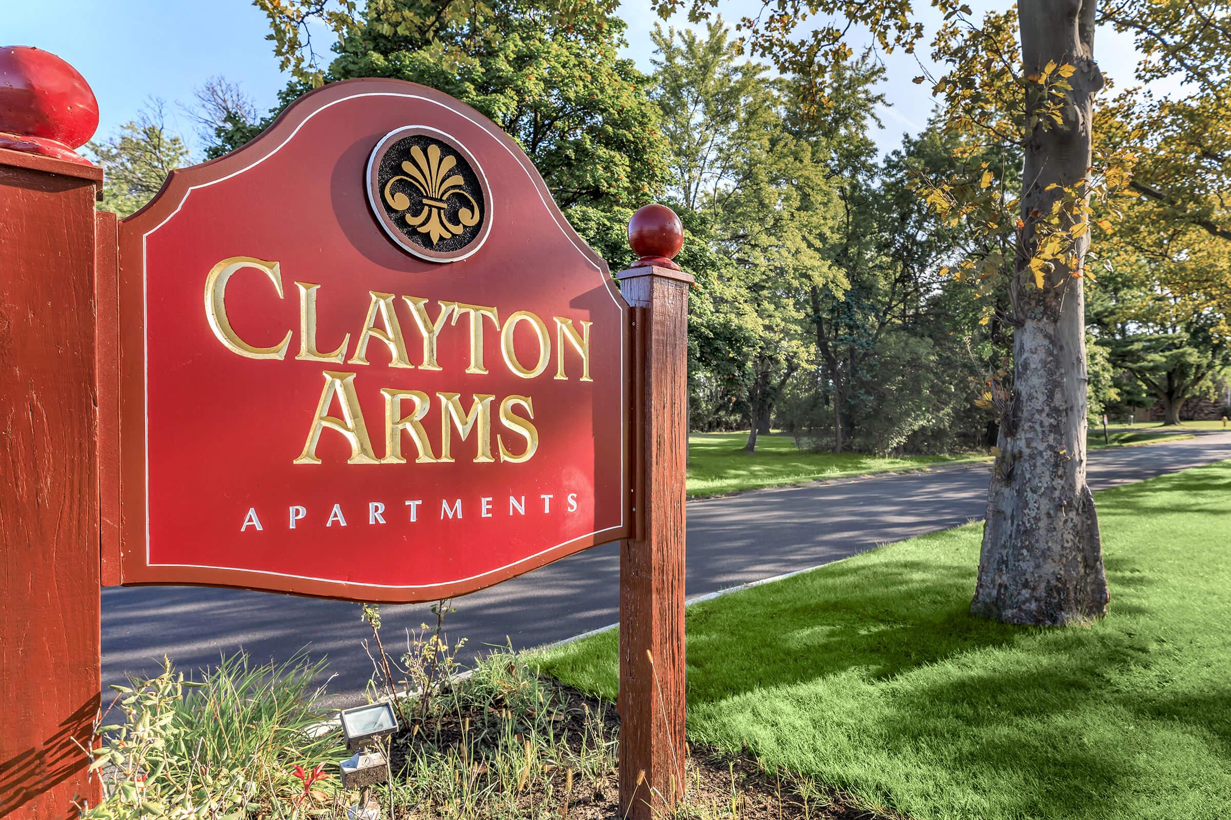 A red sign reading "Clayton Arms Apartments," with decorative elements, located beside a tree-lined road. The sign features prominent lettering and a classic design, indicating the entrance to an apartment community in a landscaped setting.