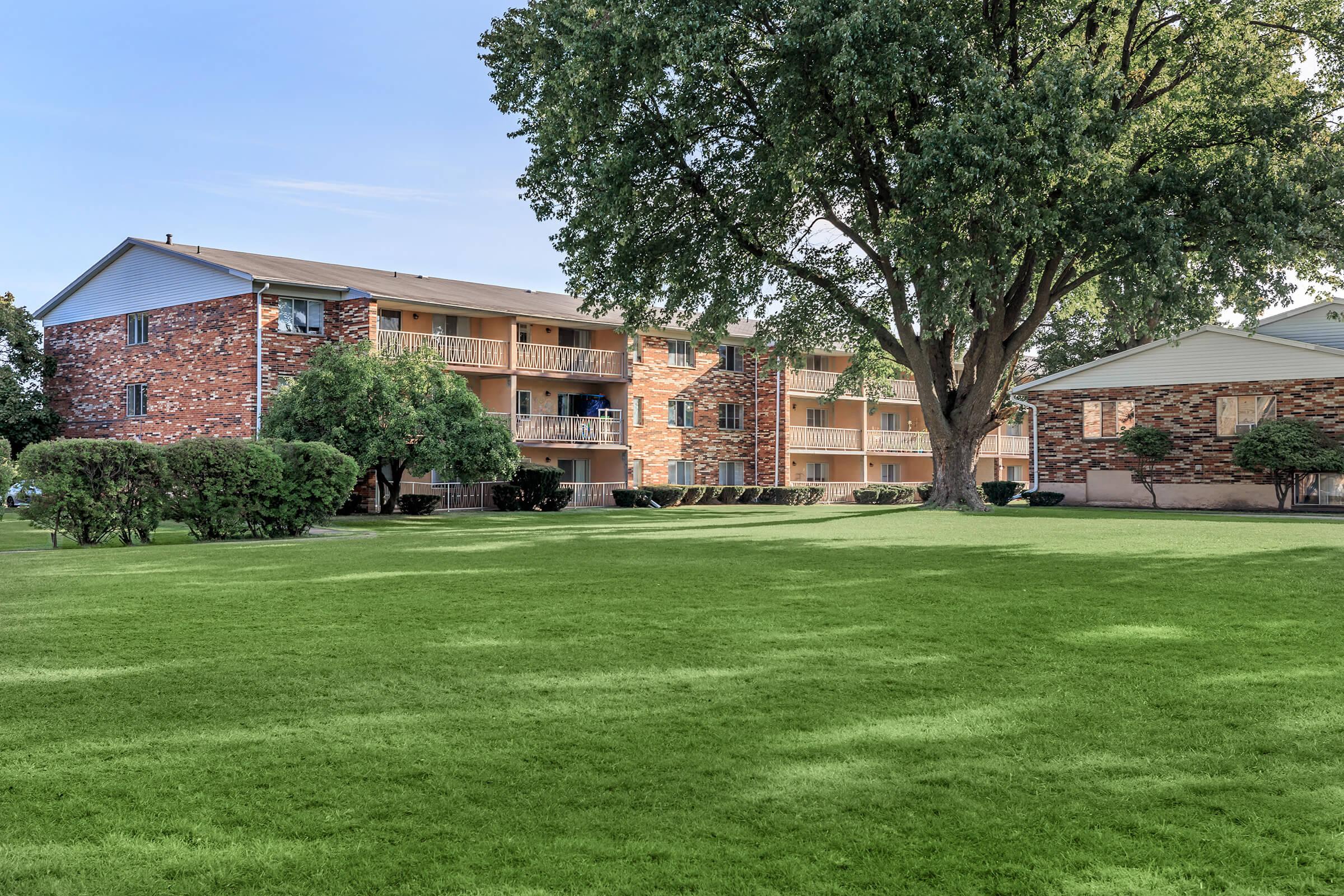 A multi-story brick apartment building surrounded by a well-manicured green lawn and trees. The building features balconies and is set against a clear blue sky, with an adjacent single-story home visible on the right.