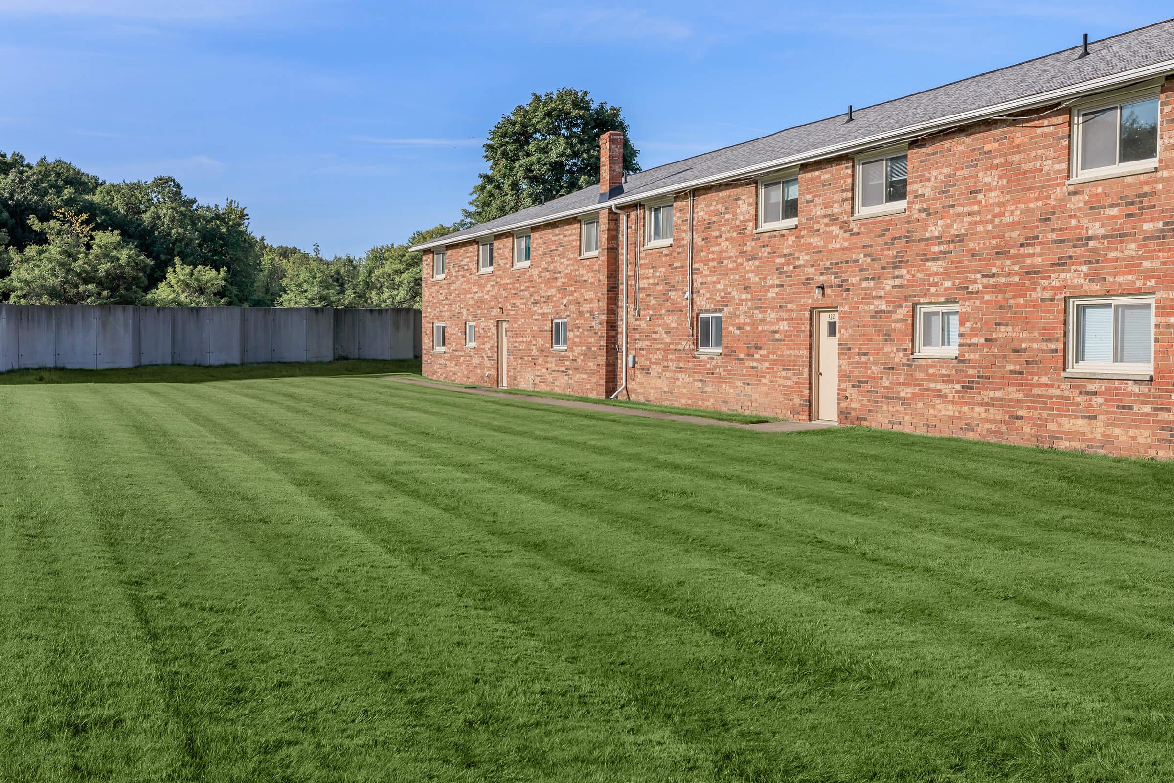 A well-maintained grassy lawn in front of a two-story brick building. The lawn is neatly mowed with visible stripes, while the building features several windows and a single door on its side. The background includes a wooden fence and trees, suggesting a peaceful residential area.