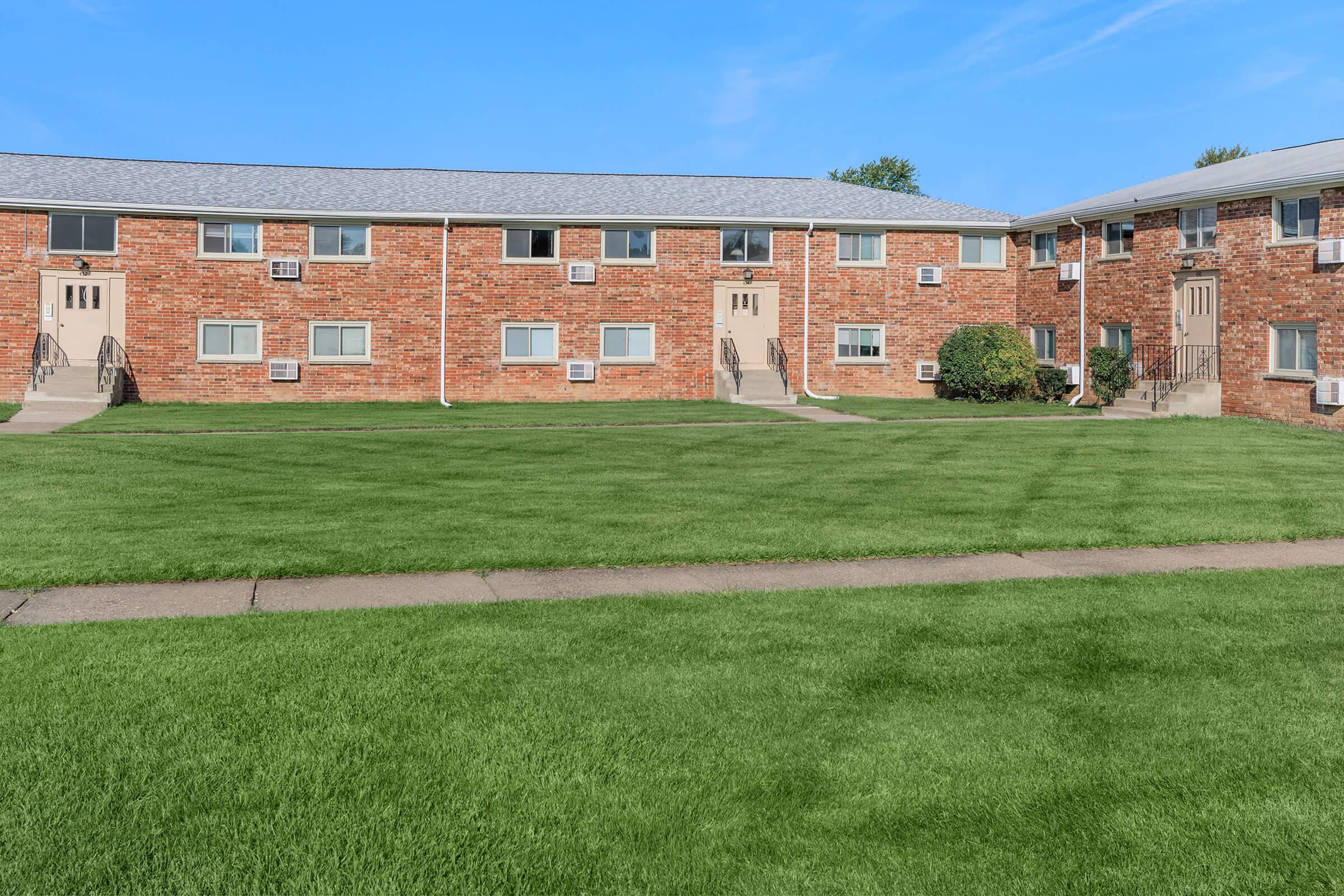 A view of a landscaped apartment community with red brick buildings. The image features two buildings facing each other across a lush green lawn with neatly trimmed grass and a concrete pathway. Each building has multiple entrances and air conditioning units in the windows. The sky is clear and blue.