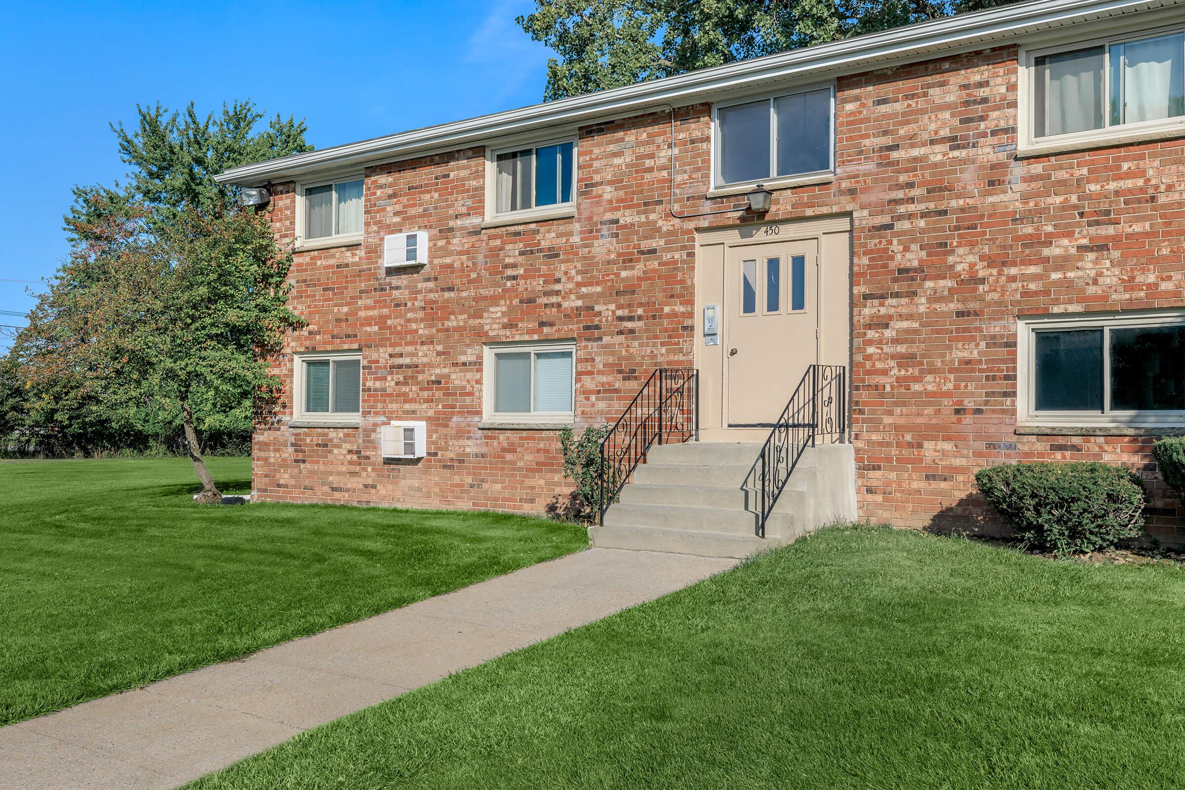 A two-story brick apartment building with multiple windows, an entrance door, and a concrete walkway. There are air conditioning units under the windows, and well-maintained green grass and shrubs surround the property. A clear blue sky is visible in the background.