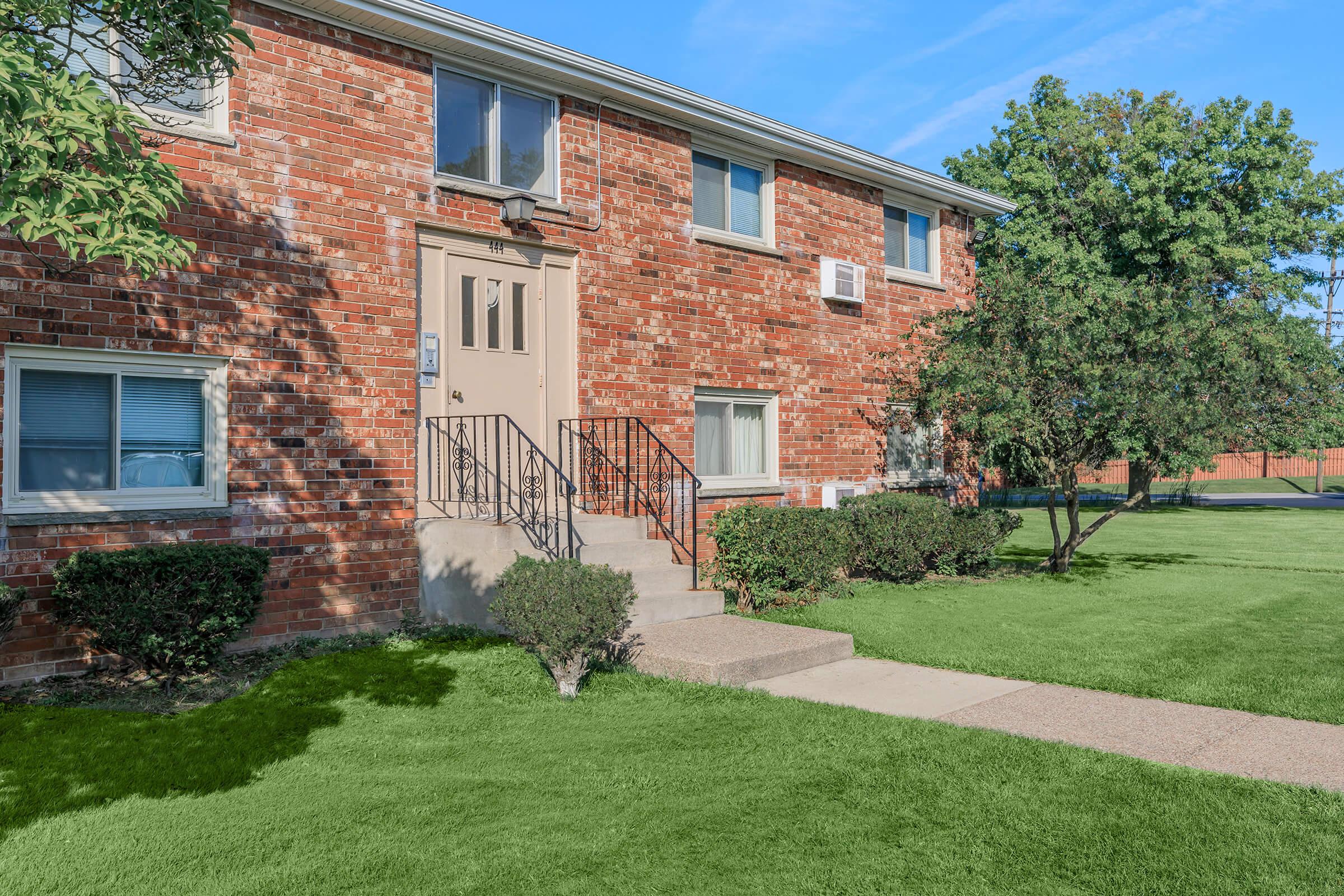 A brick apartment building with a front entrance featuring a set of stairs. Light green grass and neatly trimmed bushes surround the entrance, while trees are visible in the background under a clear blue sky.