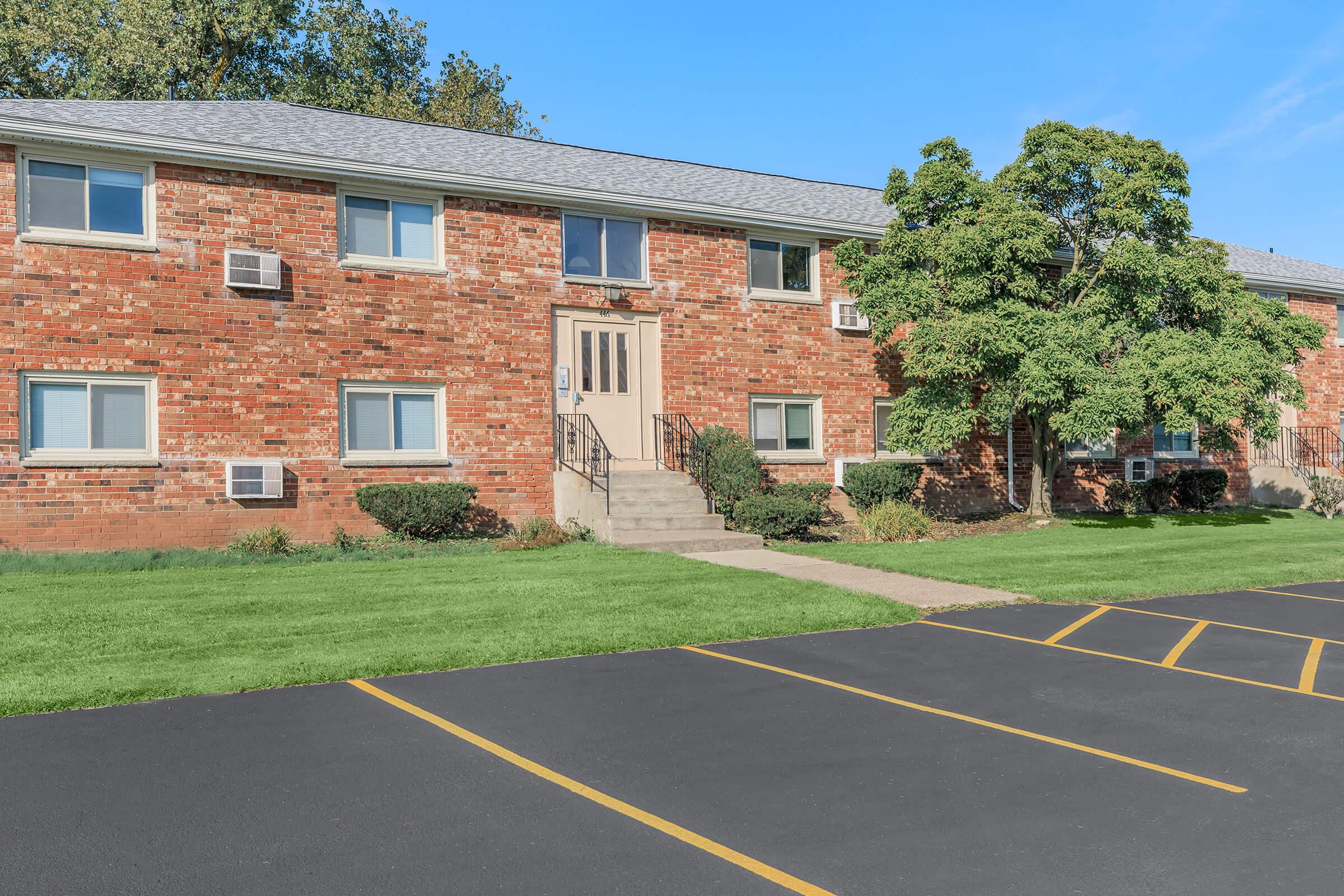 Two-story brick apartment building with multiple windows and air conditioning units. A concrete pathway leads to the main entrance, flanked by shrubs and trees. The surrounding lawn is well-maintained. A parking lot with lined spaces is visible in the foreground. Clear blue sky in the background.