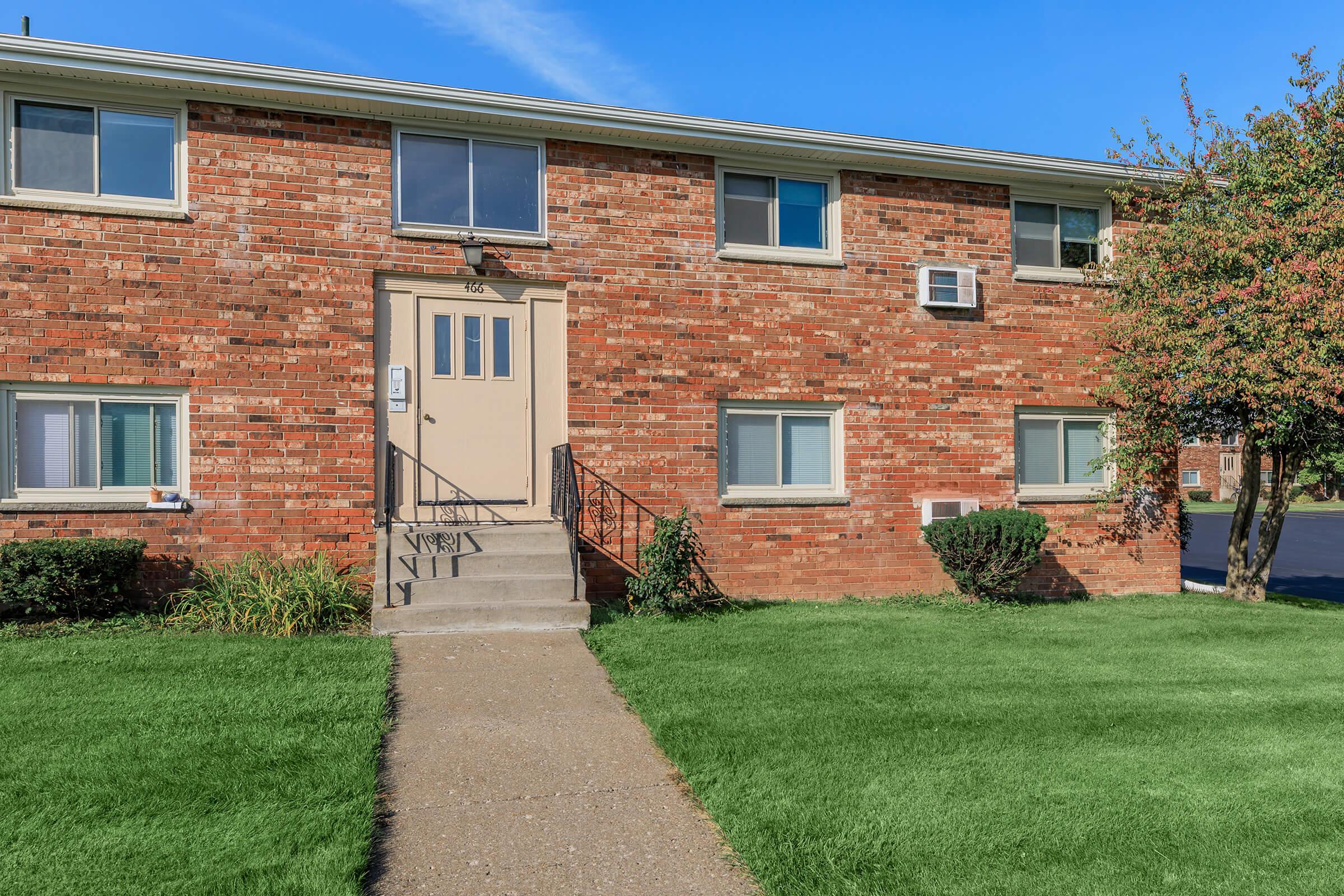 A brick apartment building with a light brown door and several windows. There is a small set of stairs leading to the entrance, and greenery including shrubs and a well-manicured lawn surrounds the building. A window air conditioning unit is installed on the side of the building. Blue sky in the background.