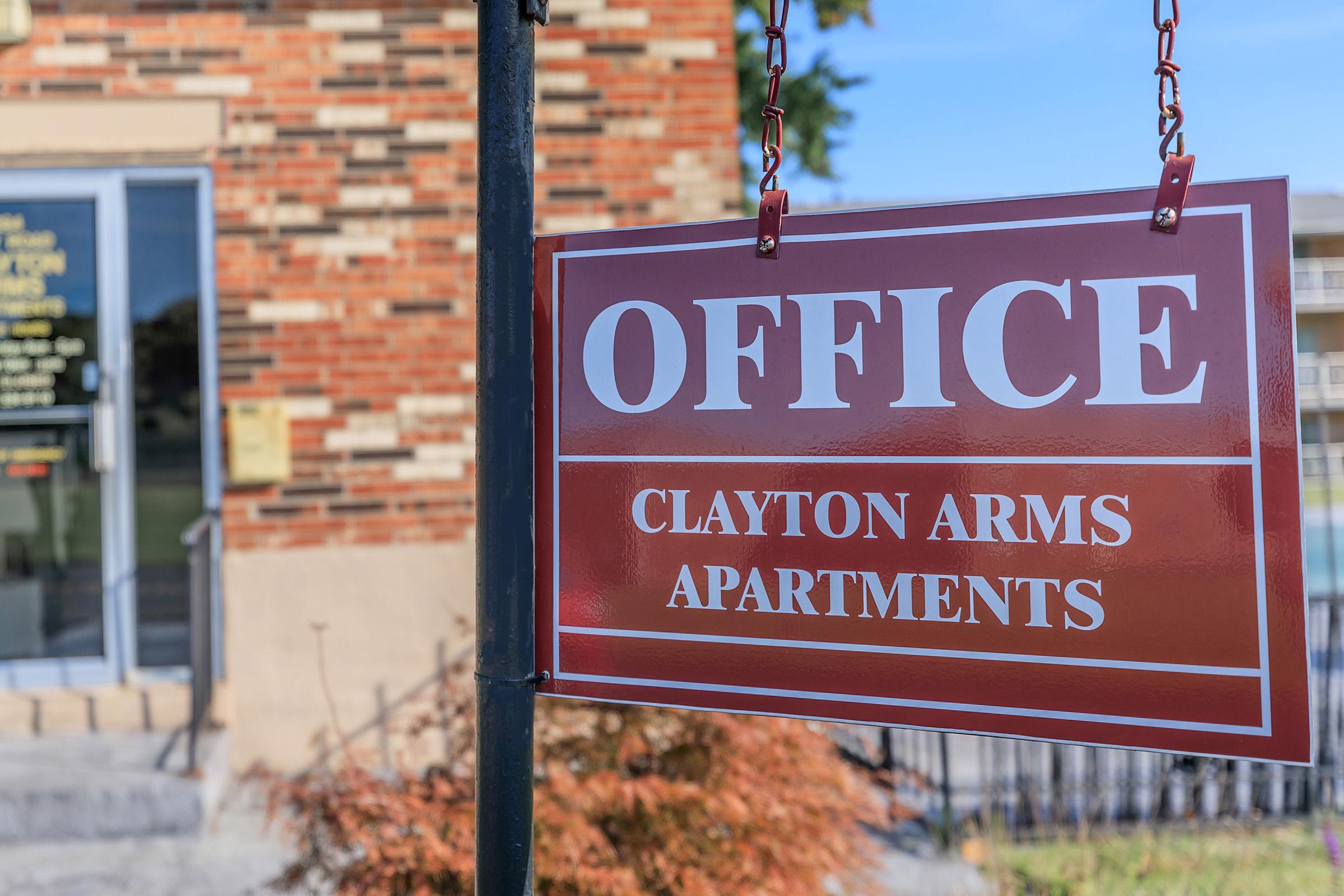 Sign indicating "OFFICE" for Clayton Arms Apartments, hanging outside a brick building. The entrance is visible in the background, with a landscaped area in front.