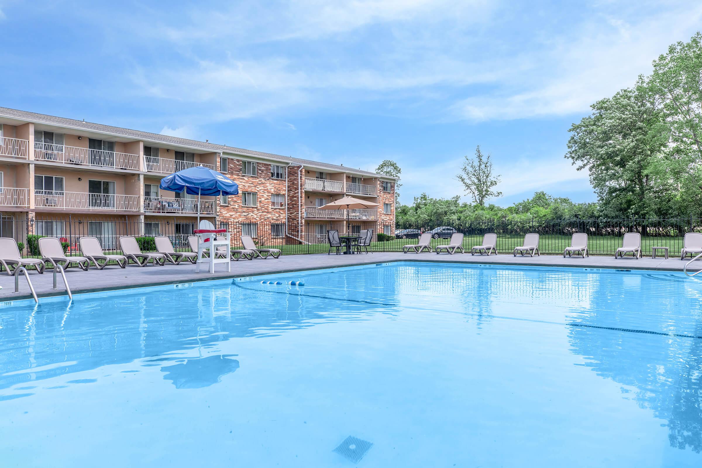 A clear swimming pool reflecting the sky, surrounded by lounge chairs and a large umbrella. In the background, a multi-story building with balconies and green trees can be seen, all under a bright blue sky. The pool area is inviting and well-maintained.