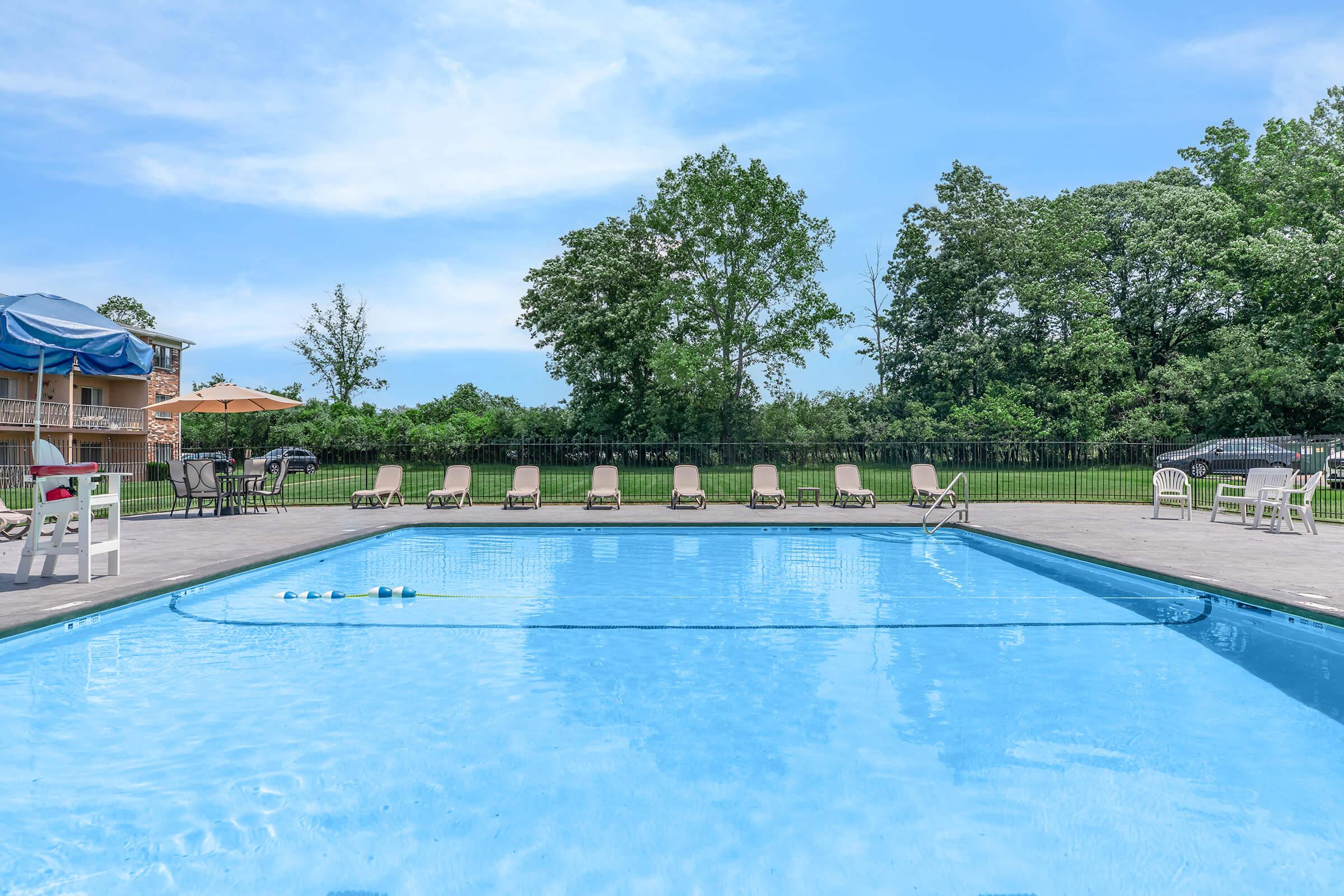 A clear swimming pool surrounded by lounge chairs and tables under a blue sky. Green trees line the background, creating a serene outdoor atmosphere. The pool features a clean, inviting surface, and the area is well-maintained, perfect for relaxation or leisure activities.