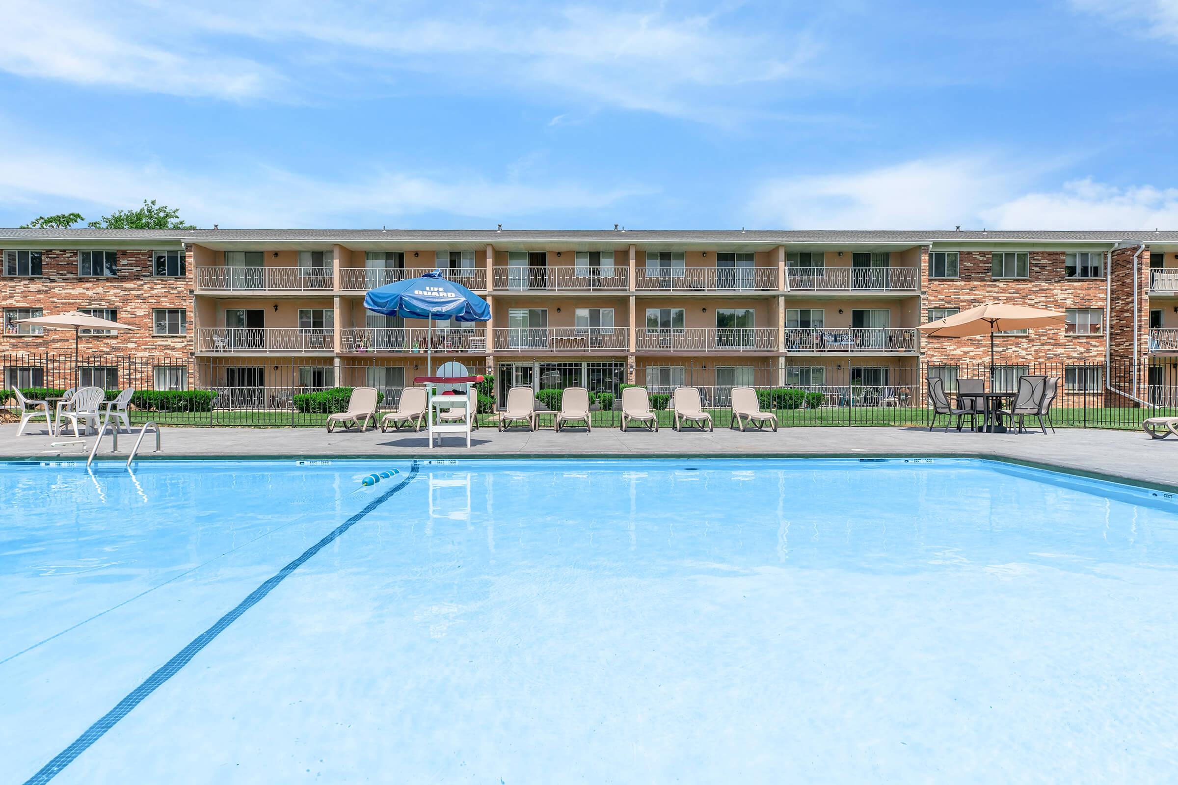 View of a swimming pool in the foreground with lounge chairs and umbrellas surrounding it, set against a backdrop of a multi-story apartment building with balconies. The sky is clear and blue, creating a bright and inviting atmosphere.