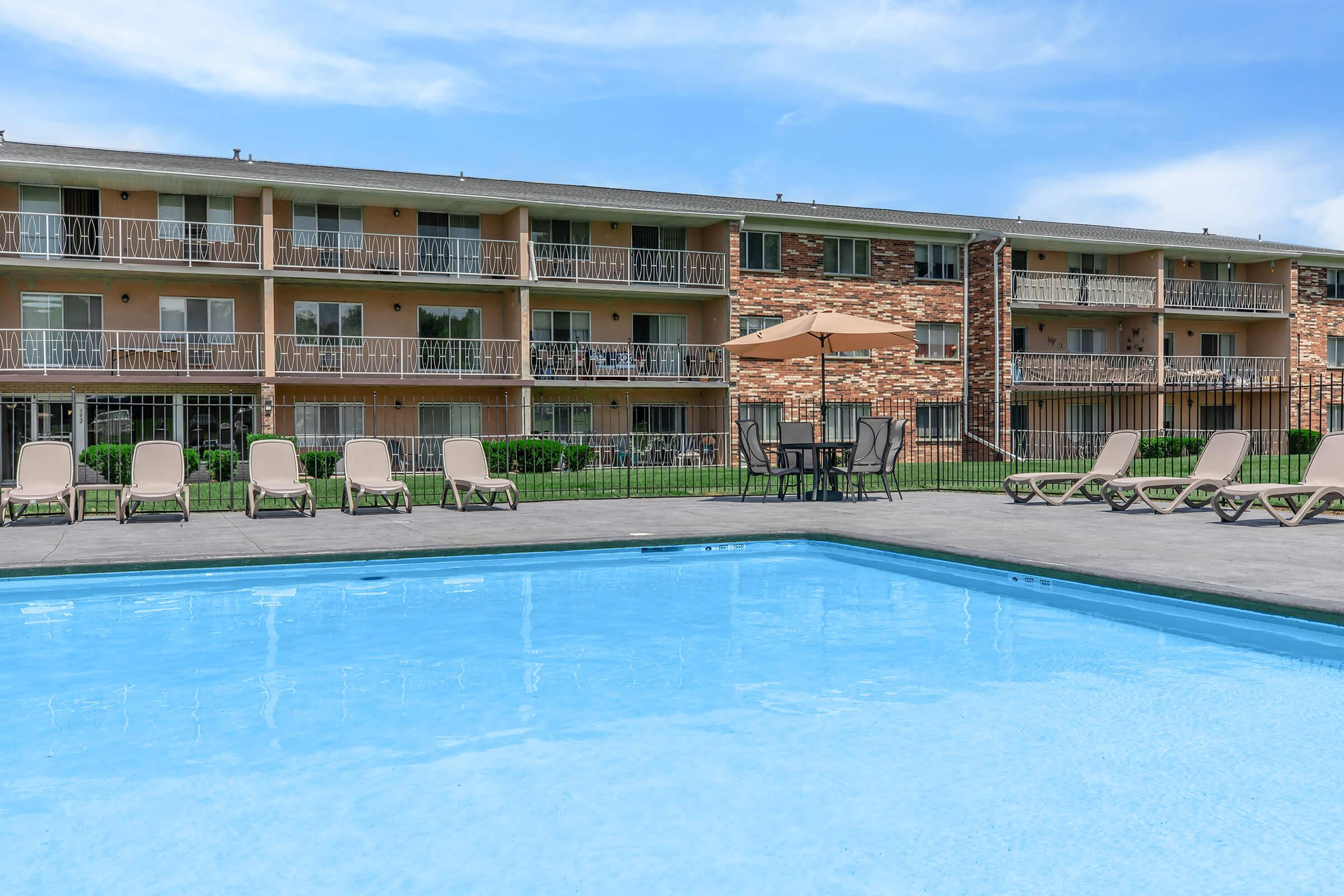 A clear swimming pool in the foreground with several lounge chairs around it. In the background, a multi-story apartment building features balconies and landscaped green spaces. The sky is bright blue with a few clouds, creating a pleasant summer atmosphere.
