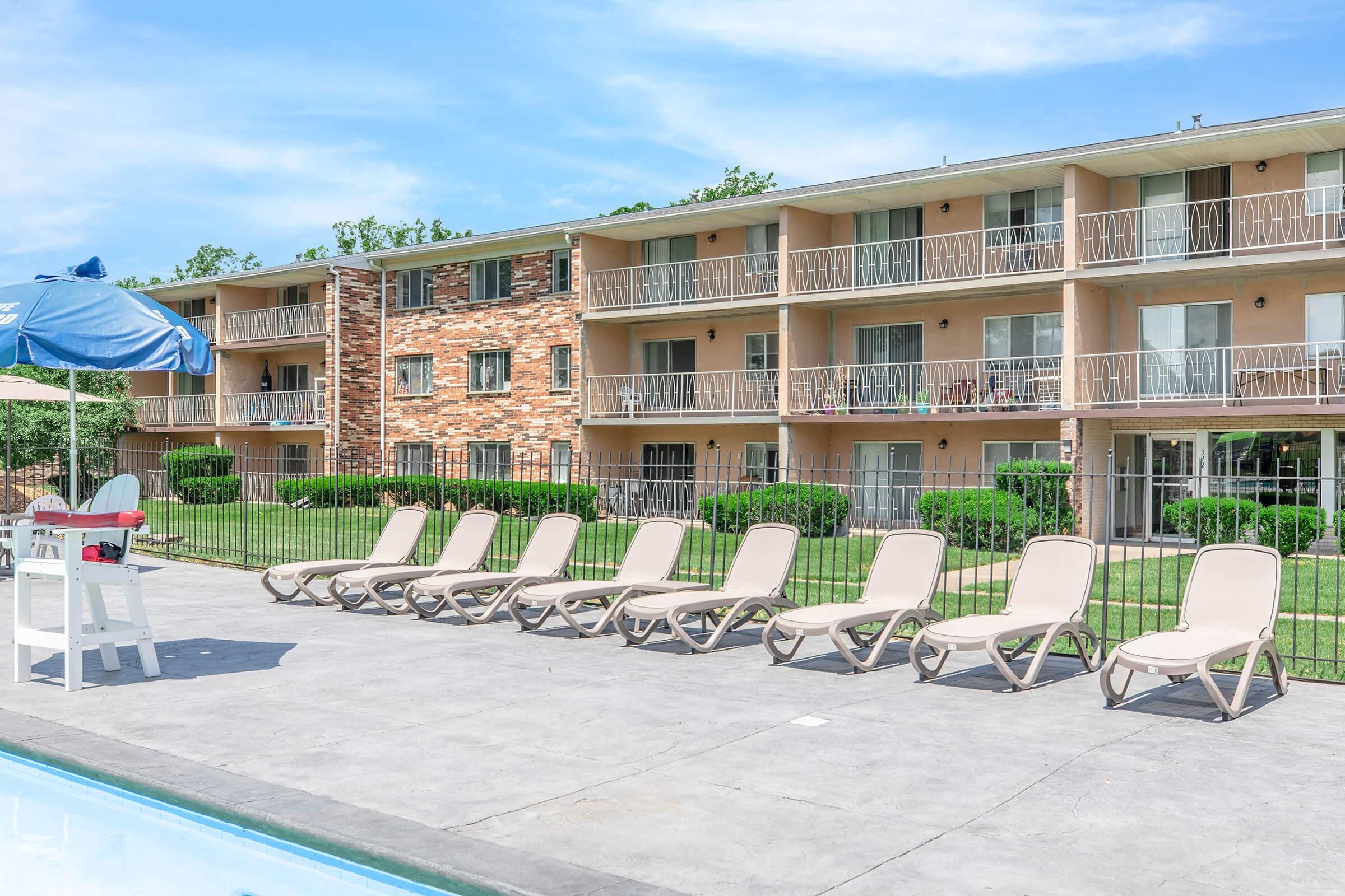 A sunny scene showing a swimming pool area with several beige lounge chairs lined up next to the water. In the background, there is a three-story apartment building with a brick facade. The surrounding area features green grass and well-maintained landscaping under a clear blue sky.