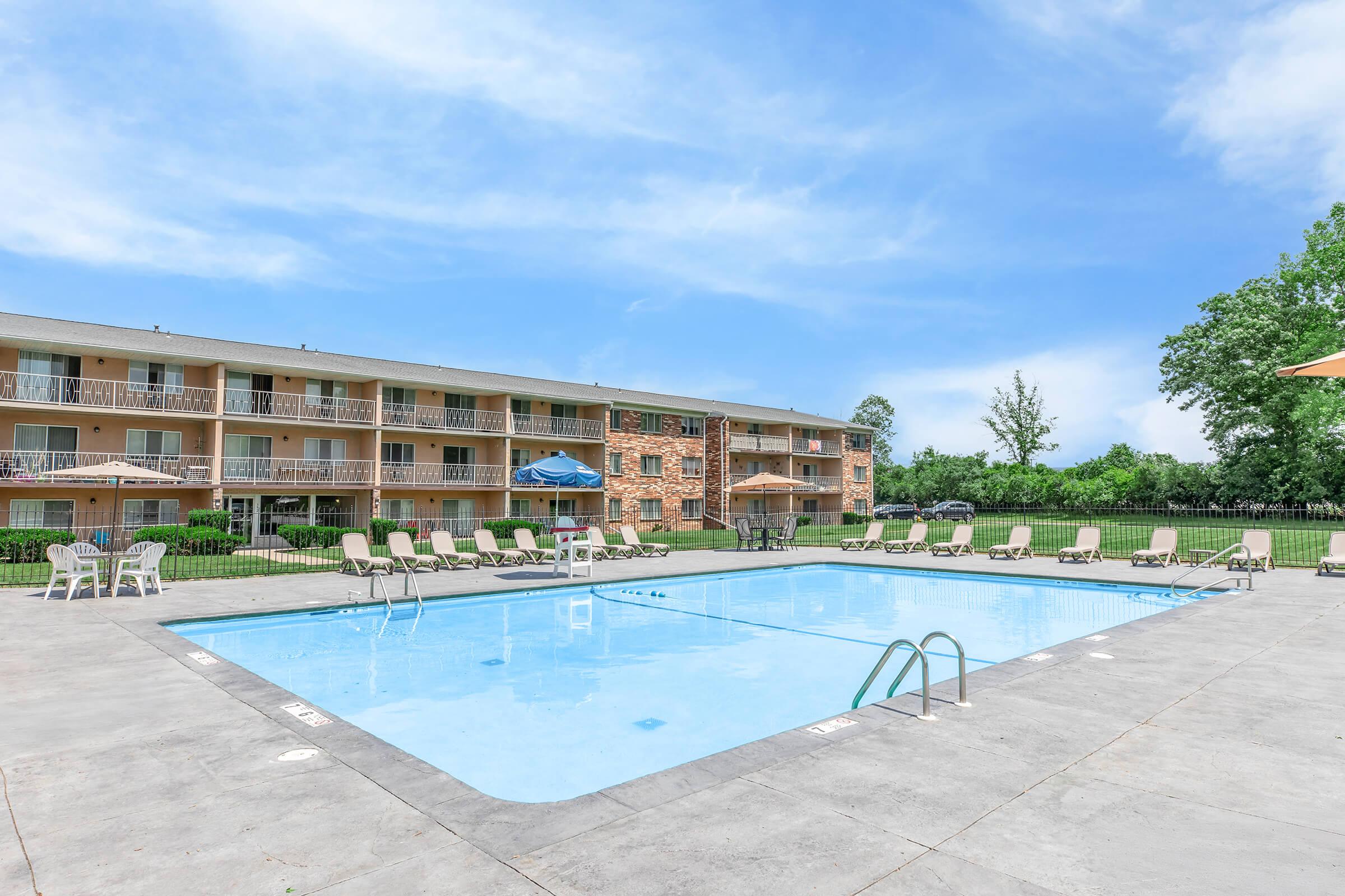 A clean, inviting outdoor swimming pool surrounded by lounge chairs. The pool area features a spacious deck, with umbrellas for shade. In the background, there are multiple residential buildings with balconies, lush green trees, and a clear blue sky overhead, creating a pleasant environment for relaxation.
