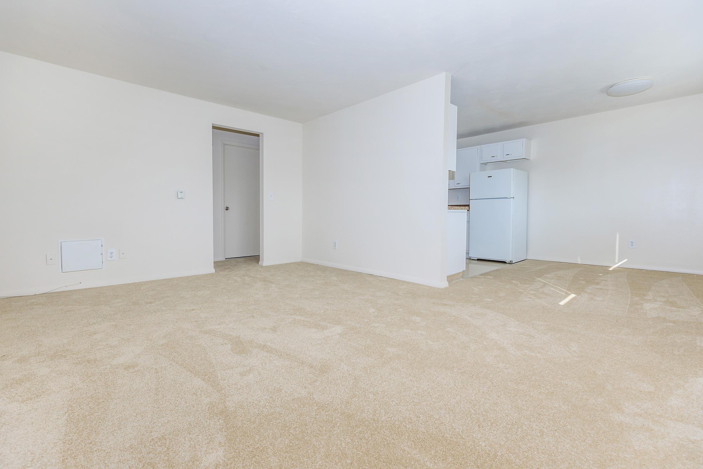 Spacious empty room with beige carpet and light-colored walls. A doorway leads to a kitchen area, featuring a white refrigerator and cabinets. Natural light illuminates the space, creating a bright and airy atmosphere.