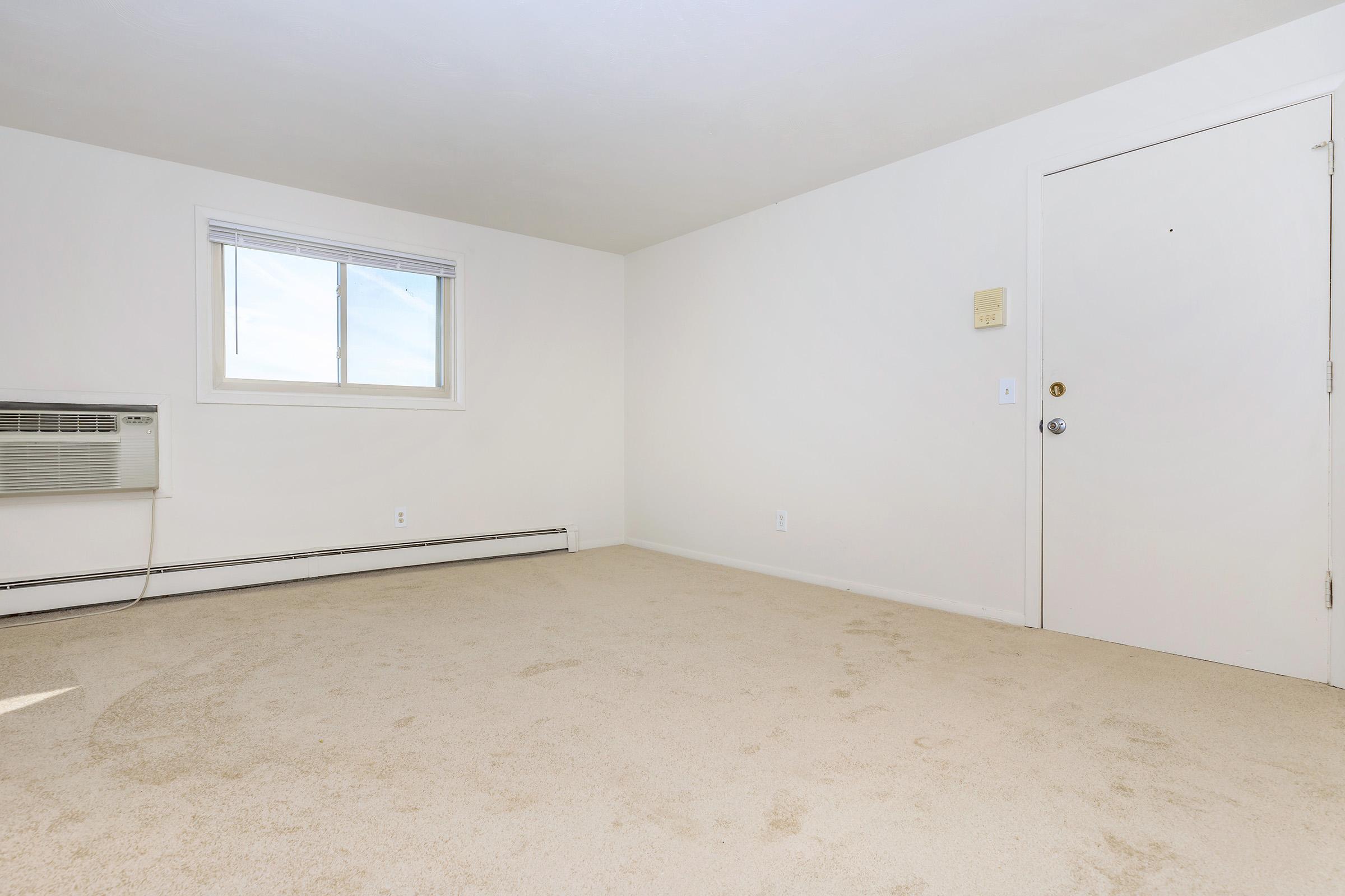 A light-colored, empty room with cream carpet and white walls. There is a small window on the left, featuring a thin white frame, and an air conditioning unit below it. The door on the right has a simple knob and is framed by white walls, creating a bright and minimalistic atmosphere.