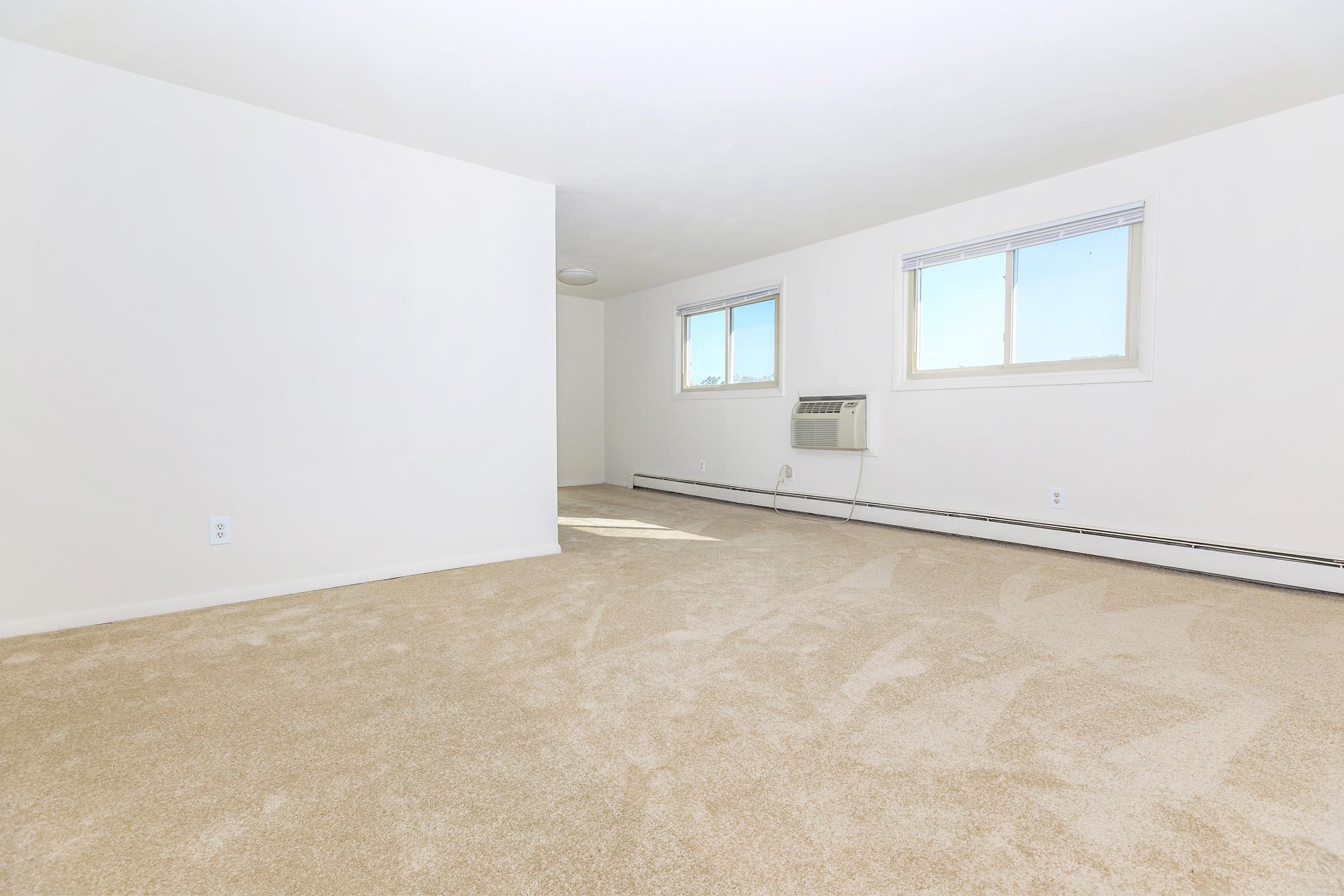 Empty room with beige carpet, white walls, and two windows letting in natural light. An air conditioning unit is mounted on the wall. The space has a clean, minimalistic appearance, suitable for various uses or furnishings.